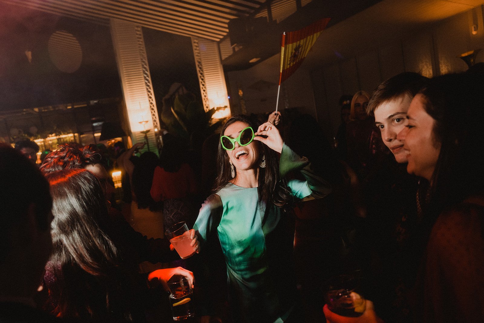 Personas disfrutando de una fiesta en un ambiente nocturno, con una mujer en el centro con gafas de sol verdes sosteniendo una bandera.