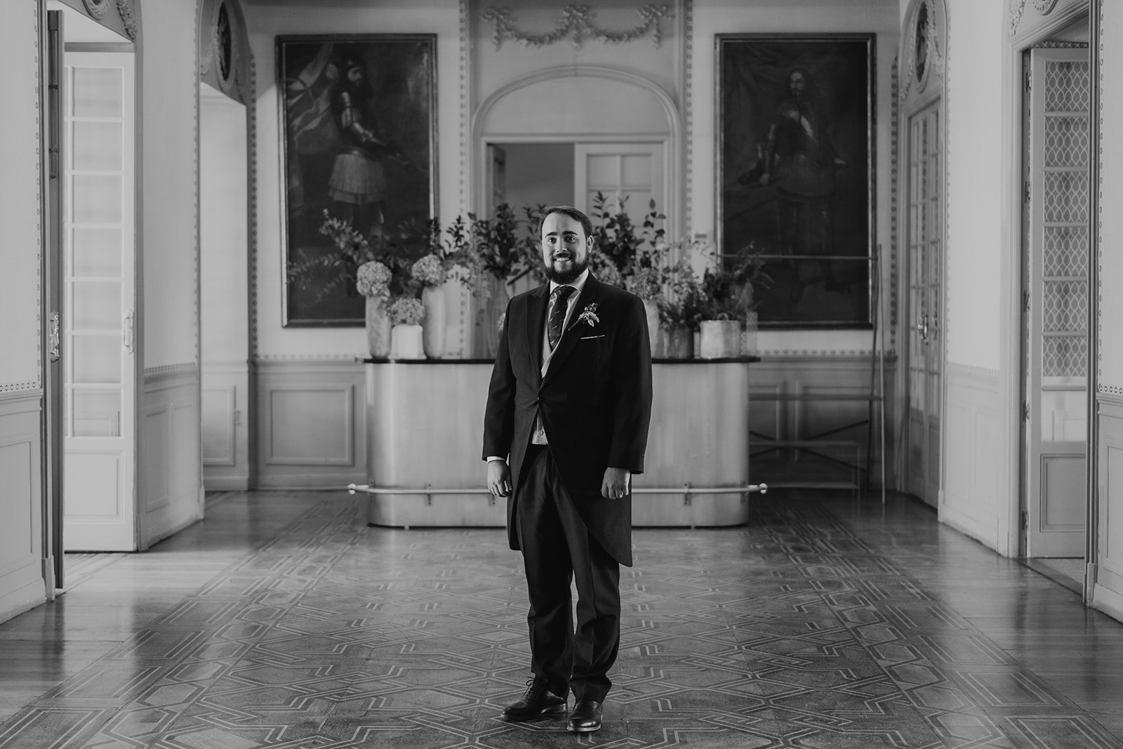 Un hombre vestido con traje formal está de pie en el centro de una sala elegante decorada con pinturas clásicas y arreglos florales.