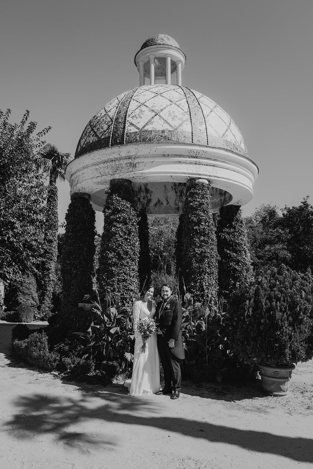 Pareja posando frente a una glorieta con techo de cúpula, rodeada de columnas cubiertas de hiedra y vegetación, en un entorno de jardín.