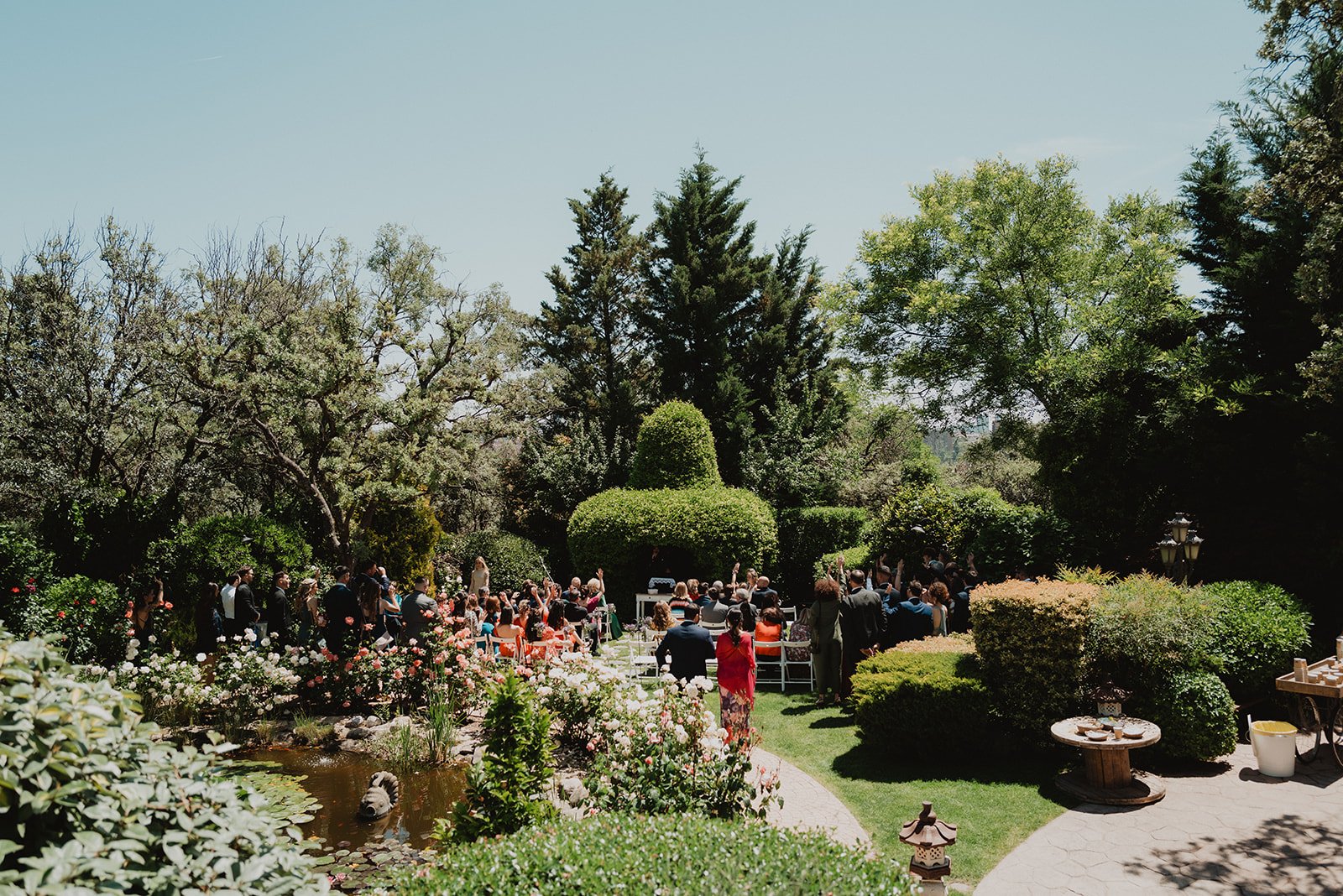 Ceremonia al aire libre en un jardín con muchas flores y un pequeño estanque, rodeada de árboles y arbustos, con personas sentadas y de pie.
