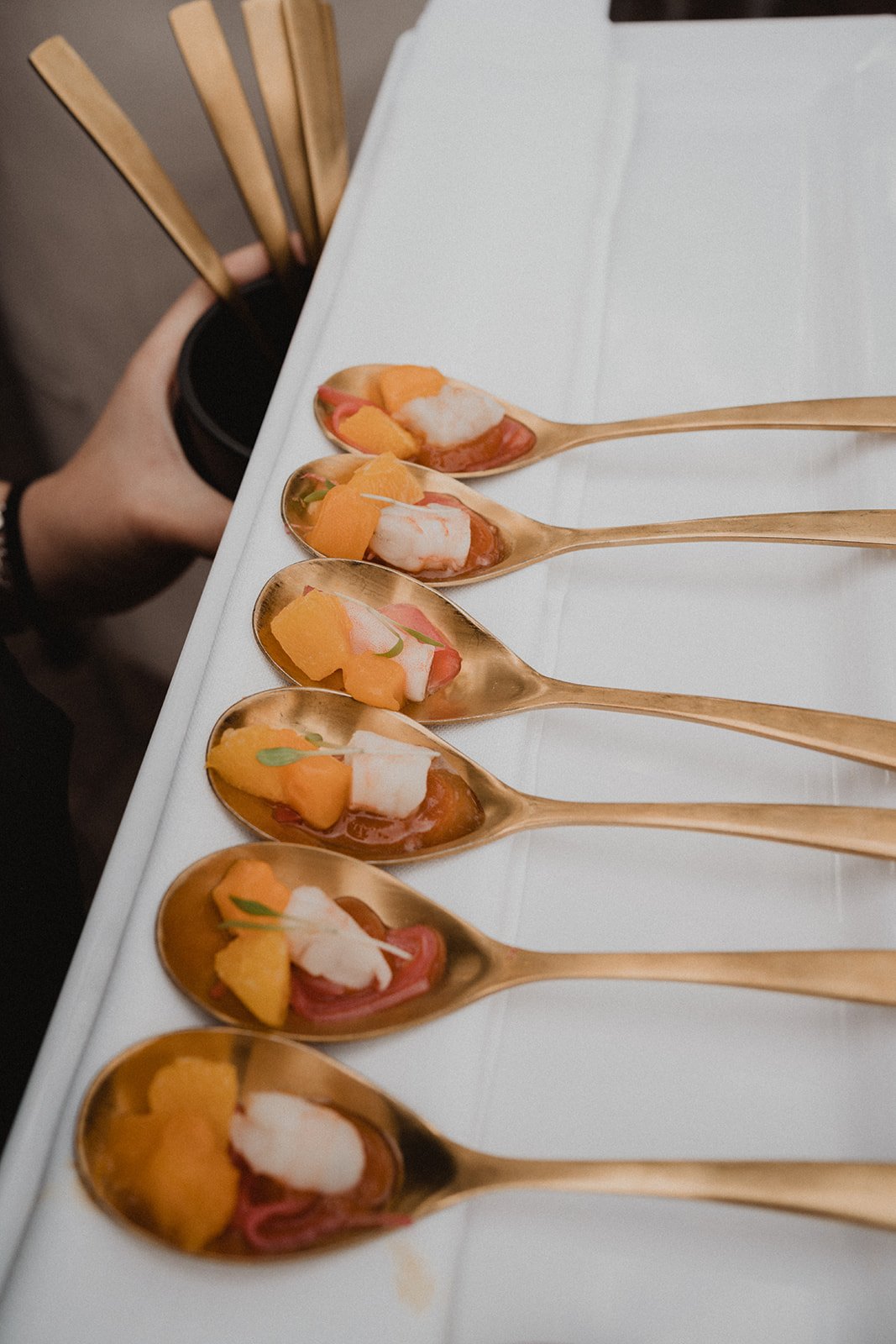 Cucharas doradas con aperitivos que incluyen mariscos y frutas, sobre una bandeja blanca.