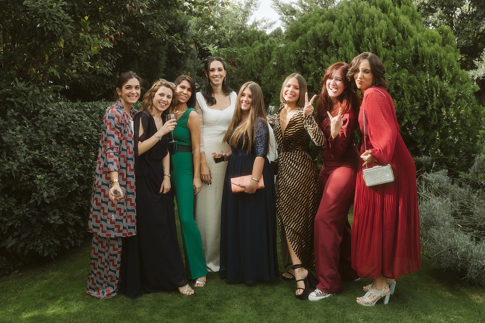 Grupo de mujeres posando en un jardín, vestidas elegantemente, algunas sosteniendo copas de bebida.