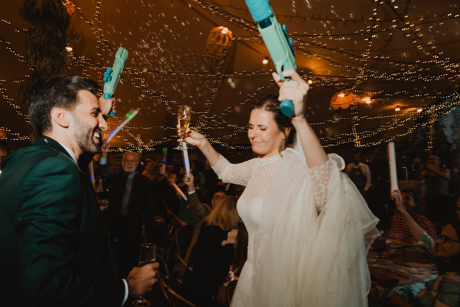 Pareja celebrando en su boda, rodeados de luces decorativas y personas festejando.