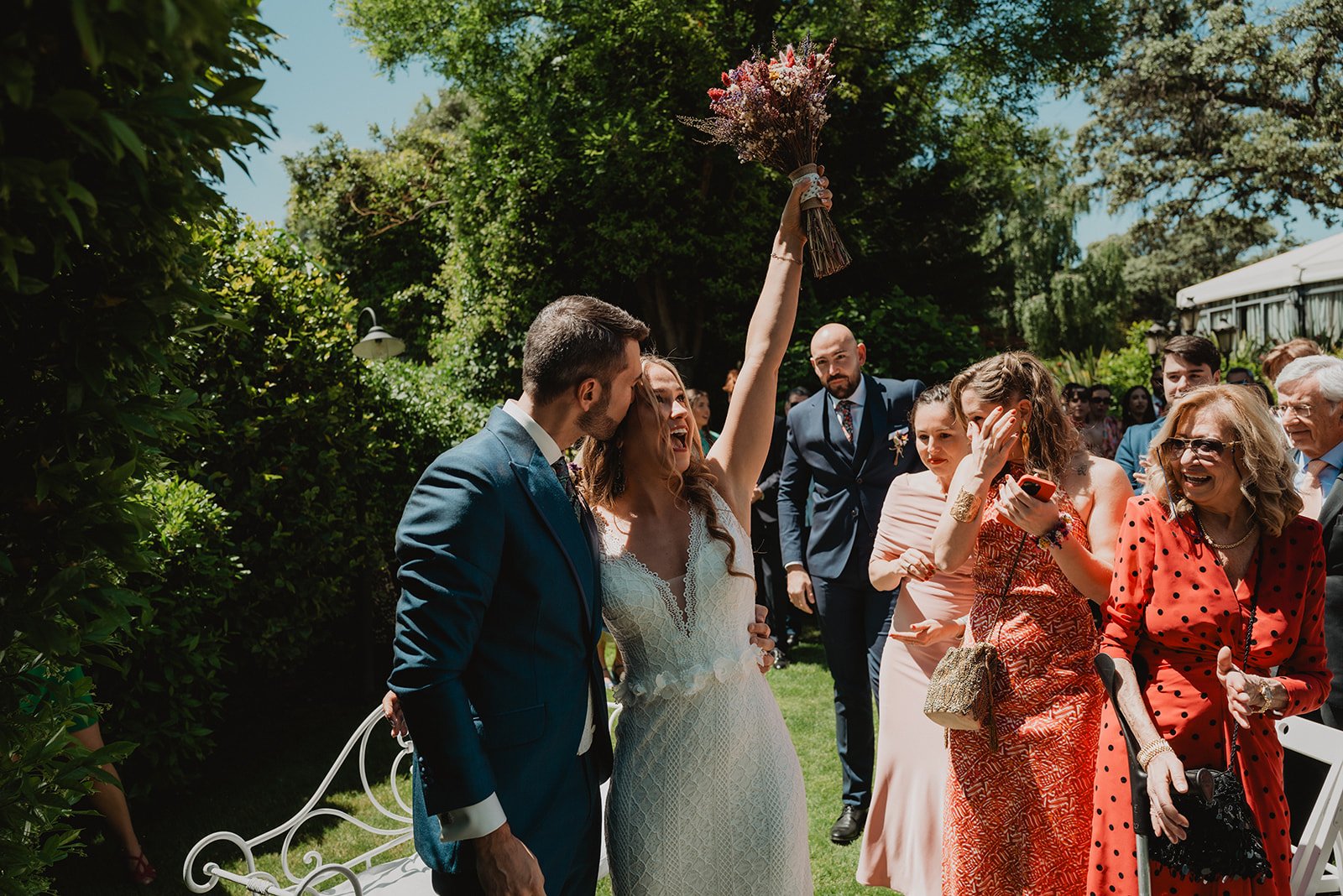 Pareja besándose en una boda con invitados celebrando al aire libre, la novia levanta un ramo de flores.