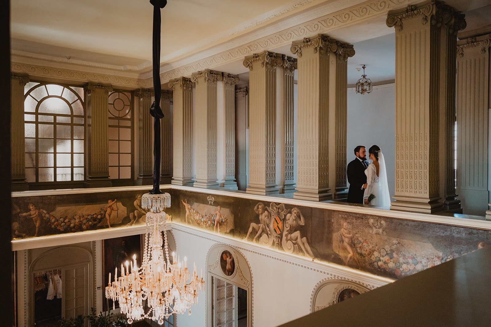 Pareja de novios en un balcón elegante con columnas y candelabro, rodeados de murales clásicos.