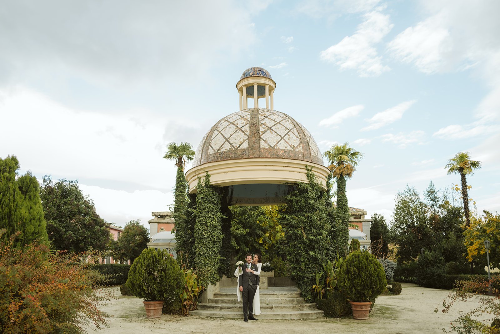 Pabellón de jardín con cúpula, cubierto de hiedra, rodeado de plantas y palmeras, con una pareja posando en la entrada.