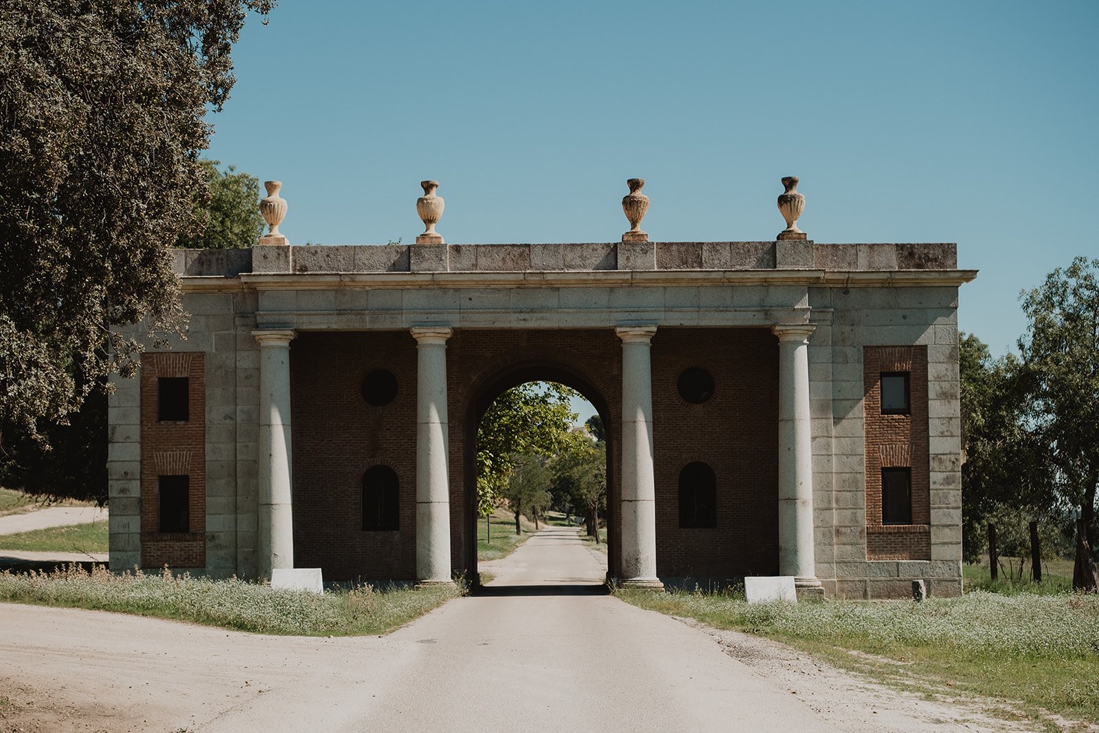 Puerta histórica de piedra con columnas y esculturas en la parte superior, rodeada de árboles y un camino pavimentado.