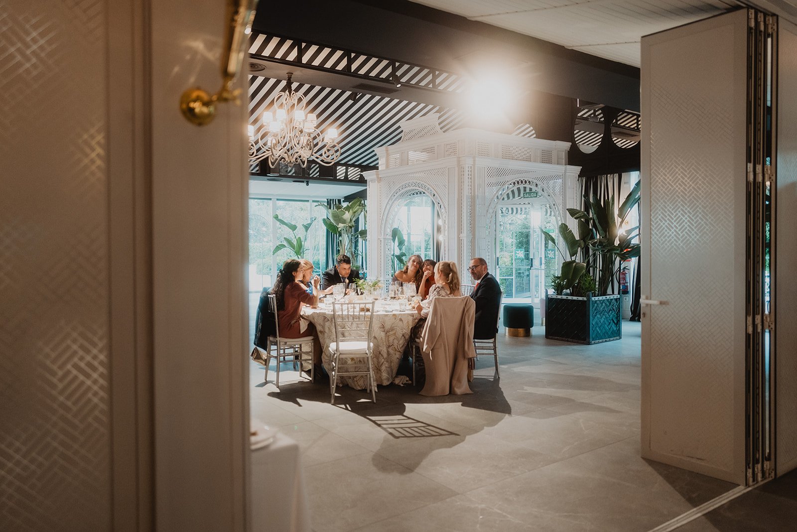 Evento social en un elegante salón interior, con personas sentadas alrededor de una mesa redonda, decorada con un mantel blanco, bajo una araña de luces.
