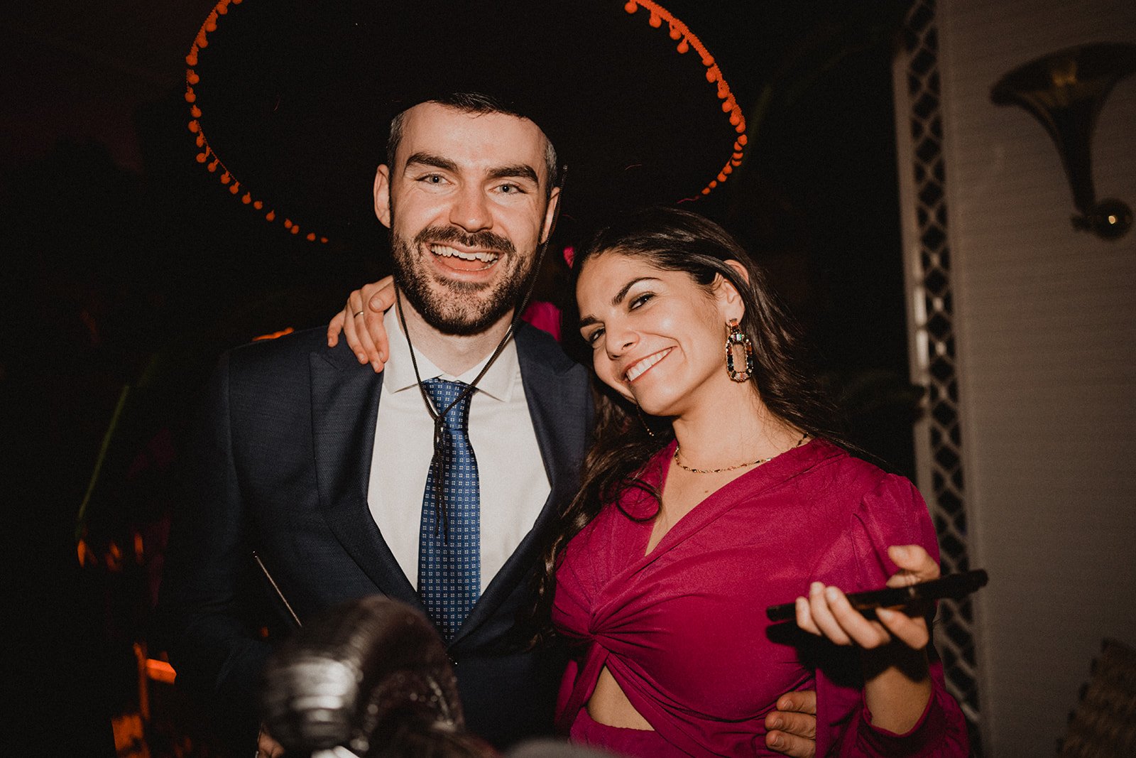 Una pareja sonriendo; el hombre lleva un sombrero charro y traje formal, mientras que la mujer usa un vestido rosa y sostiene un teléfono móvil.