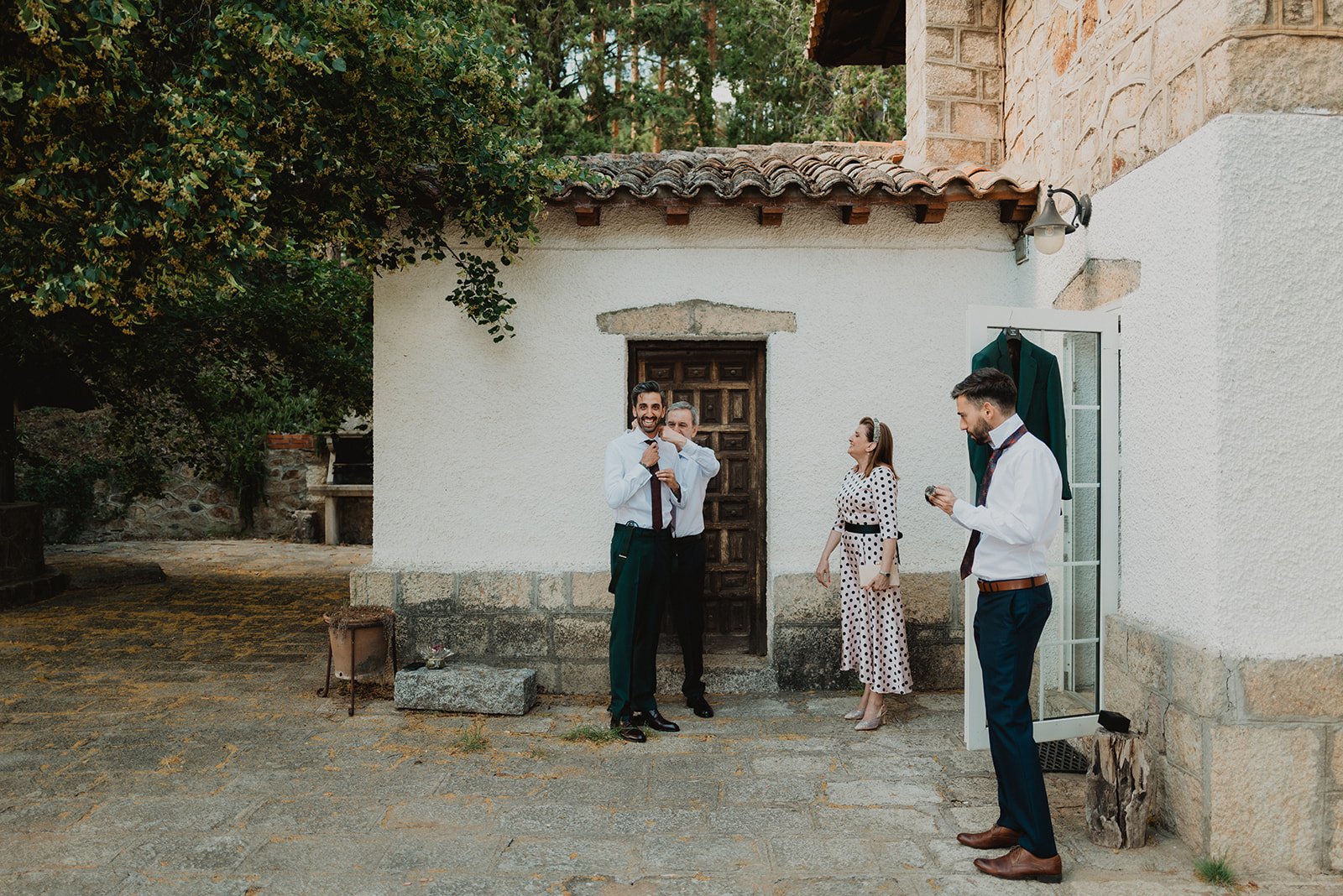 Cuatro personas vestidas formalmente afuera de una casa de piedra blanca, con árboles y vegetación alrededor. Un hombre ajusta su corbata mientras otros observan.
