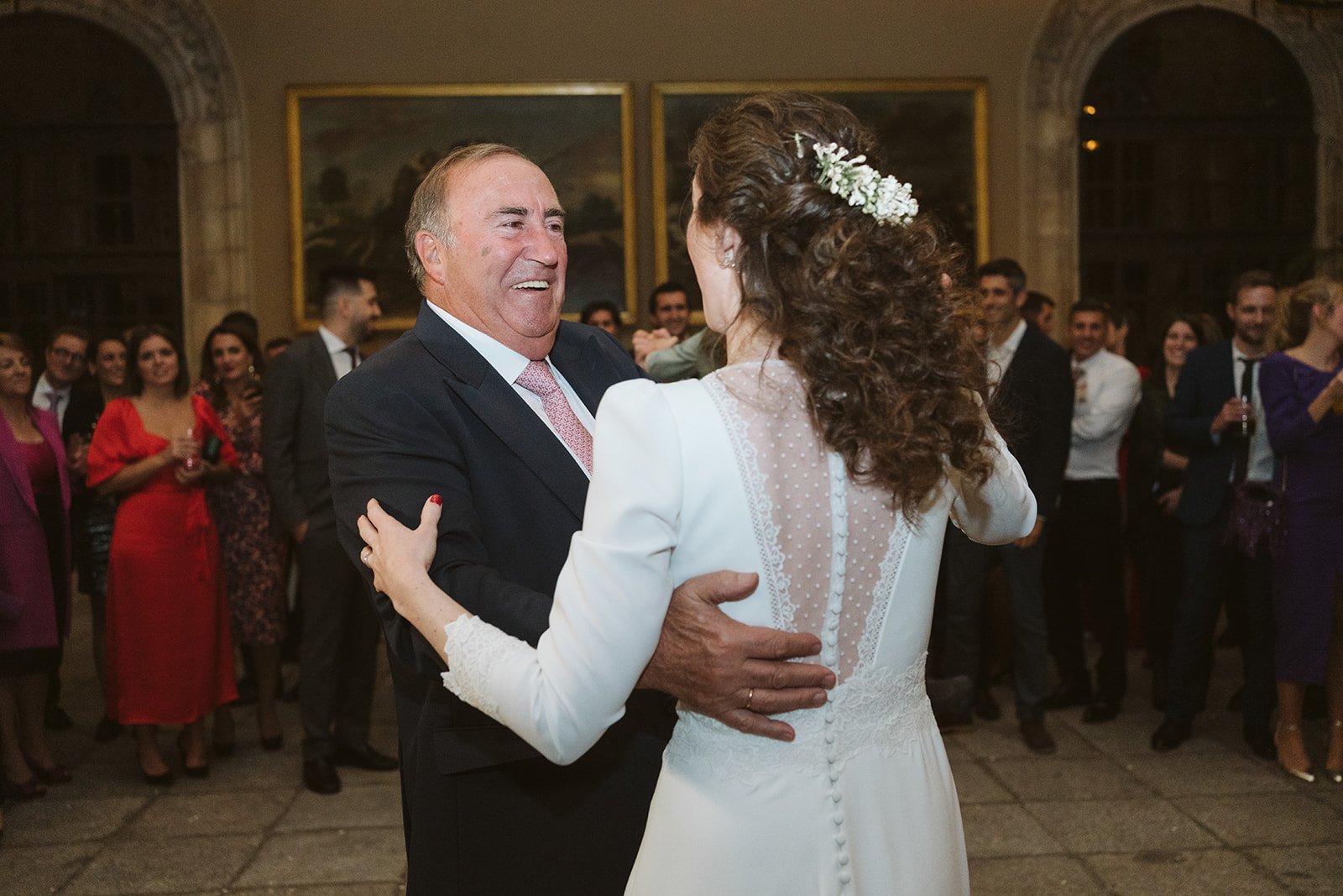 Un hombre mayor y una mujer joven bailando en una boda, rodeados de invitados bien vestidos.