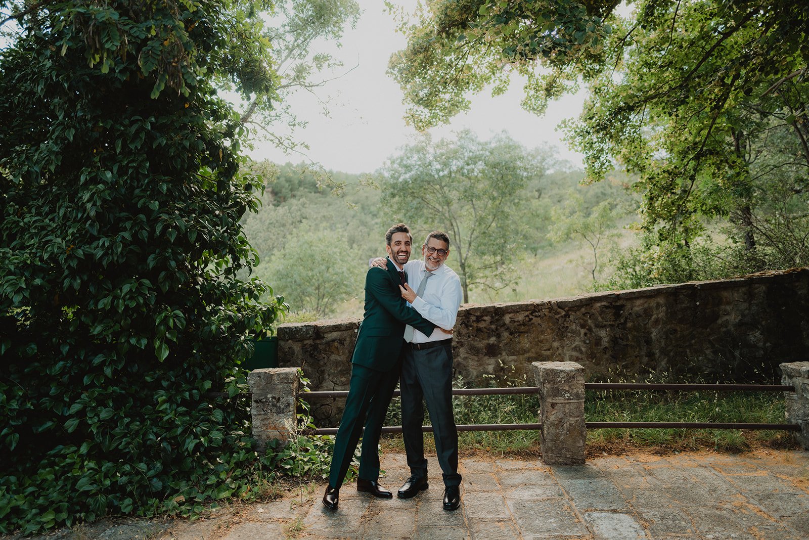 Dos hombres abrazados en un jardín con vegetación abundante y árboles, sonrientes. Ambos llevan trajes formales; uno en traje verde oscuro y el otro en camisa blanca y pantalón negro. Escenario natural al fondo.