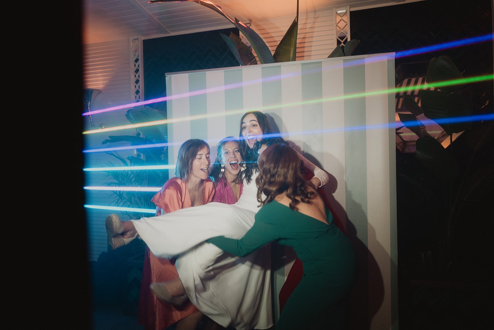Cuatro mujeres sonriendo y levantando a una amiga vestida de blanco en una fiesta, con luces de colores pasando por el frente.