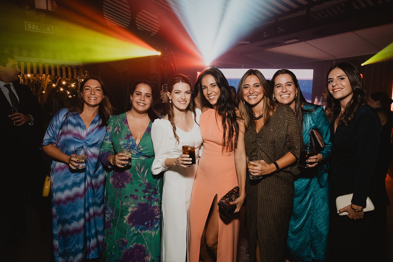 Un grupo de mujeres sonrientes posando para una foto en una fiesta, con luces de colores en el fondo.