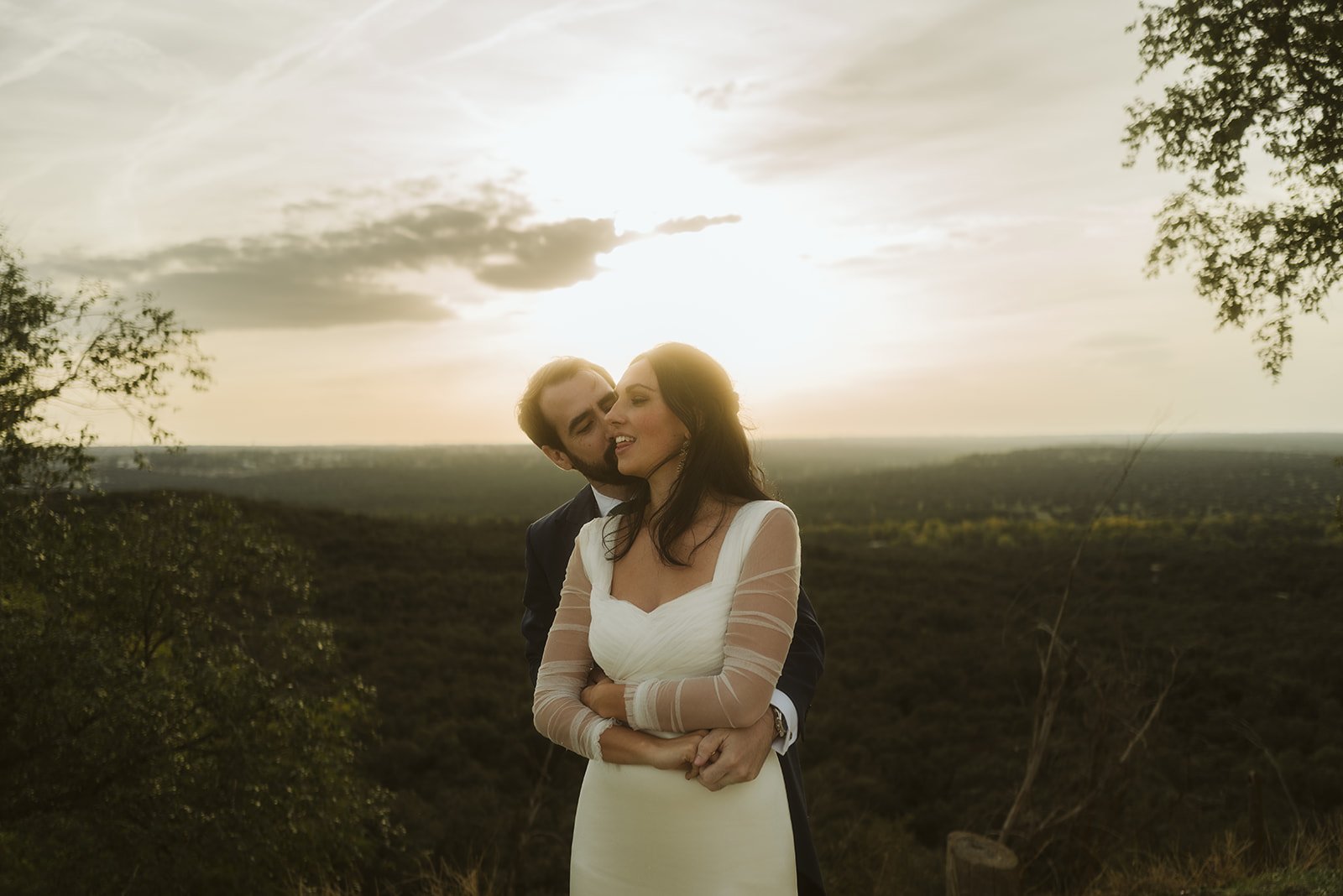 Pareja abrazada al atardecer en un paisaje natural, con cielo nublado y árboles en el fondo.