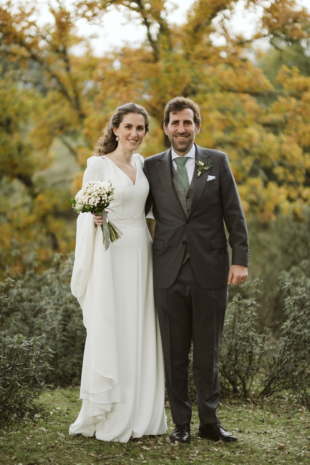 Pareja de recién casados posando en un entorno natural con fondo de árboles otoñales. La novia lleva un vestido blanco y sostiene un ramo de flores, mientras el novio viste un traje oscuro con corbata verde.