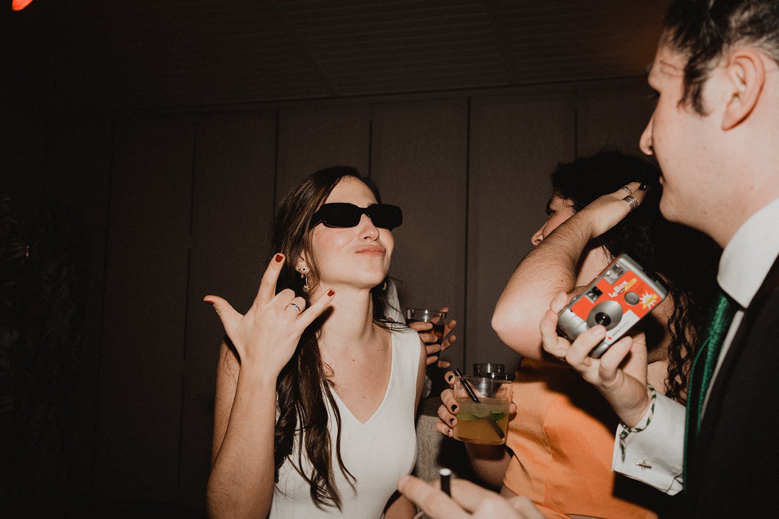 Personas divirtiéndose en una fiesta, con gafas de sol, sosteniendo bebidas y una cámara desechable.