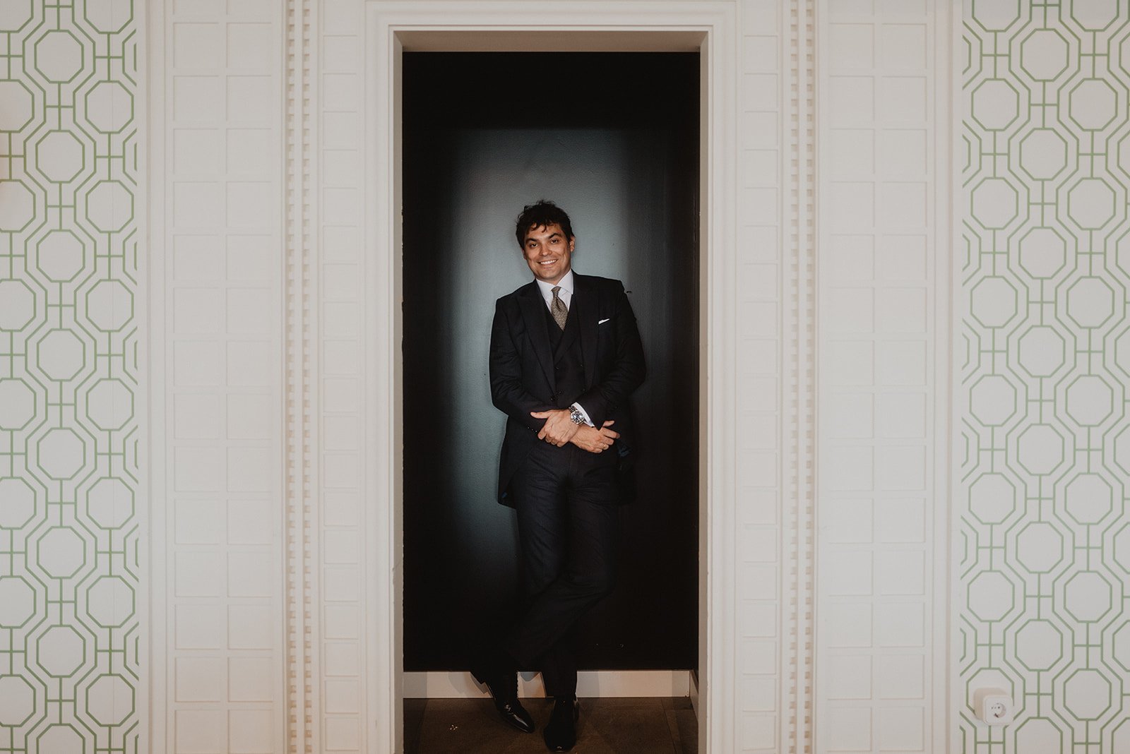 Hombre en traje oscuro posando en un marco de puerta, con fondo de pared con diseño geométrico.