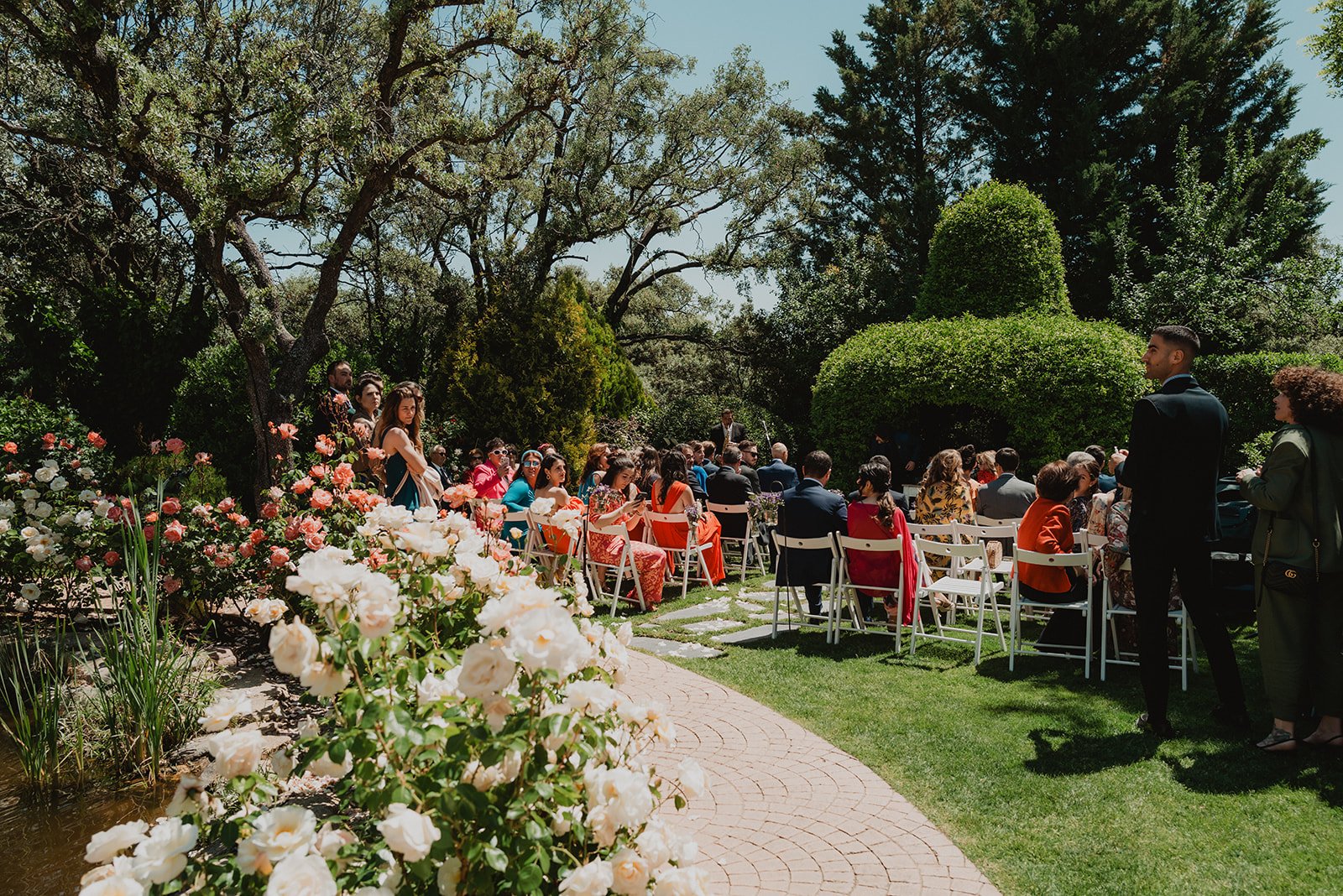 Ceremonia al aire libre con invitados sentados en sillas blancas, rodeados de jardines y flores, bajo árboles grandes.