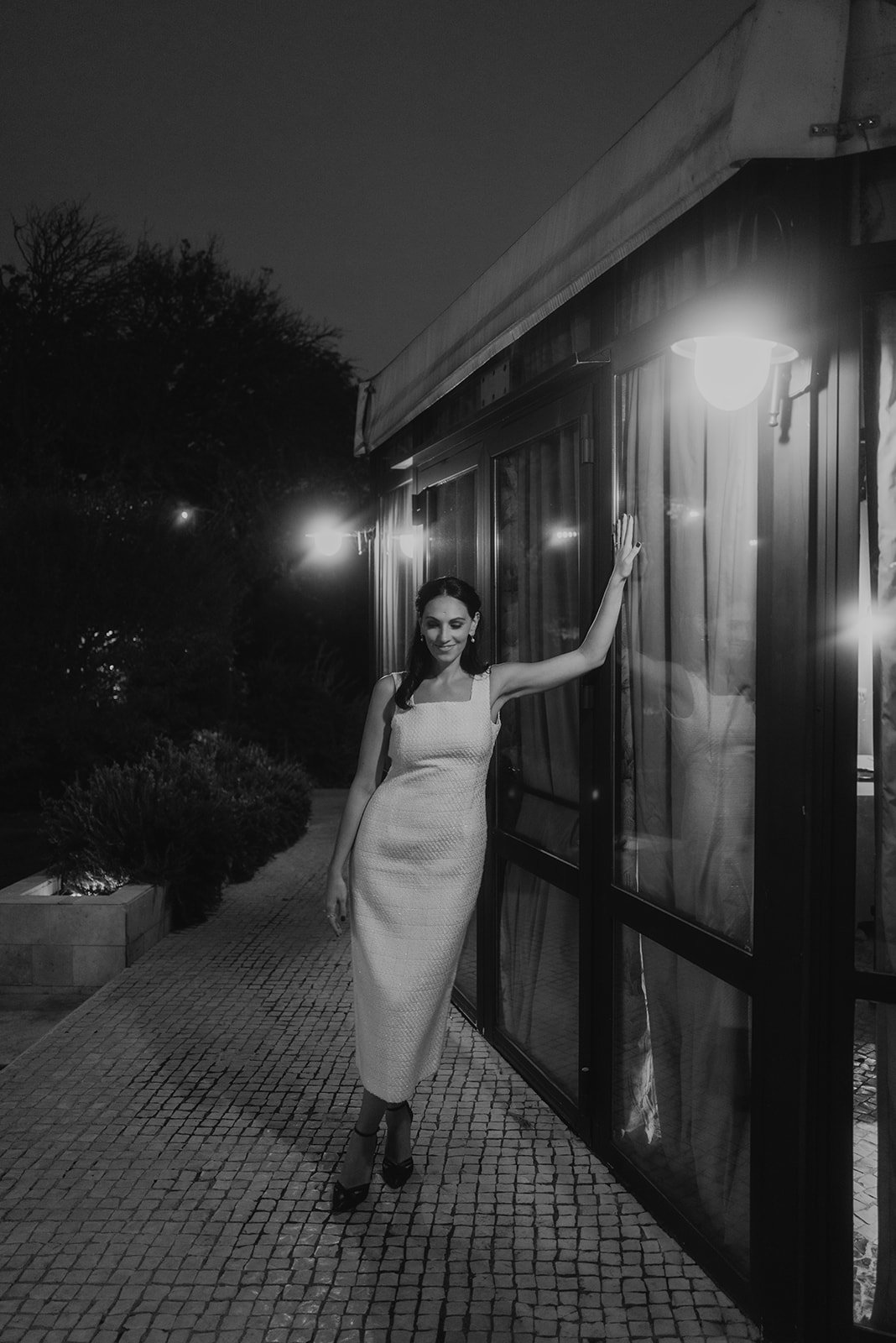 Mujer con vestido largo blanco, posando junto a una pared con ventanas en un entorno al aire libre, durante la noche. La escena está iluminada por lámparas.