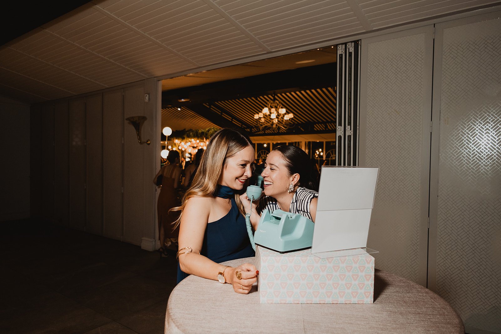 Dos mujeres felices usando un teléfono antiguo en un evento, sobre una mesa redonda con un mantel claro, en un lugar decorado con luces y lámparas al fondo.