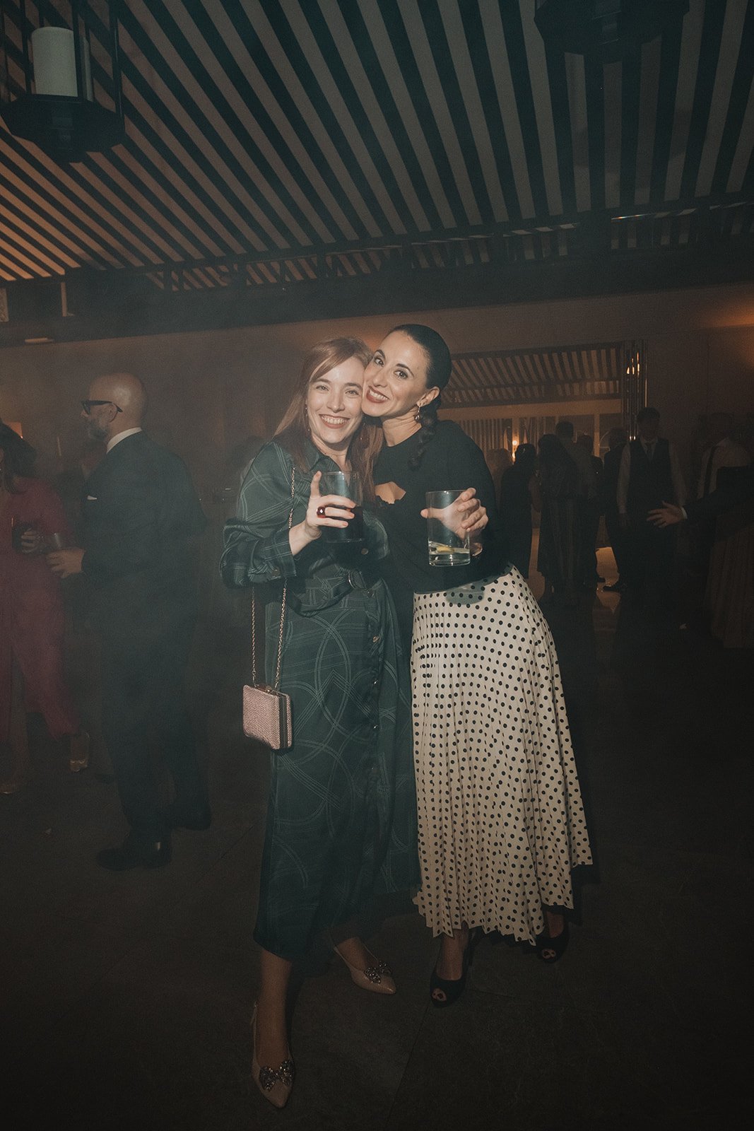 Dos mujeres sonrientes en una fiesta sosteniendo bebidas, en un ambiente con luces tenues y personas al fondo.