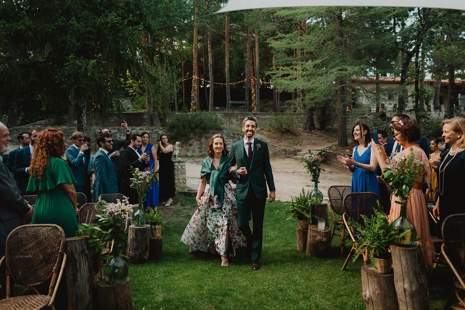 Una pareja camina por el pasillo en una boda al aire libre, rodeada de invitados que aplauden. El entorno es natural con árboles y decoración floral.