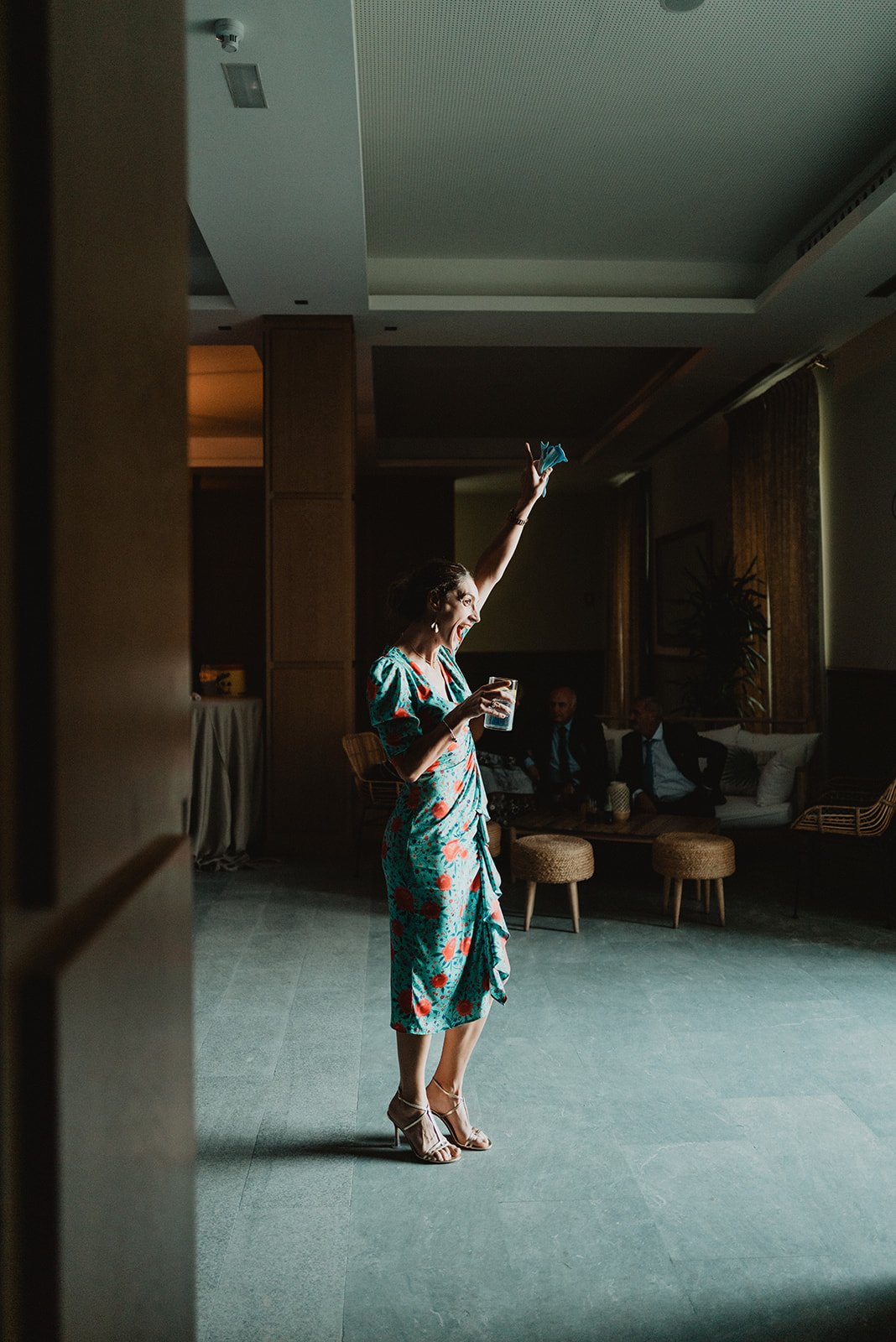 Mujer levantando el brazo con un vaso en la mano, vistiendo un vestido verde con estampado, en una habitación con poca luz y muebles de mimbre en el fondo.