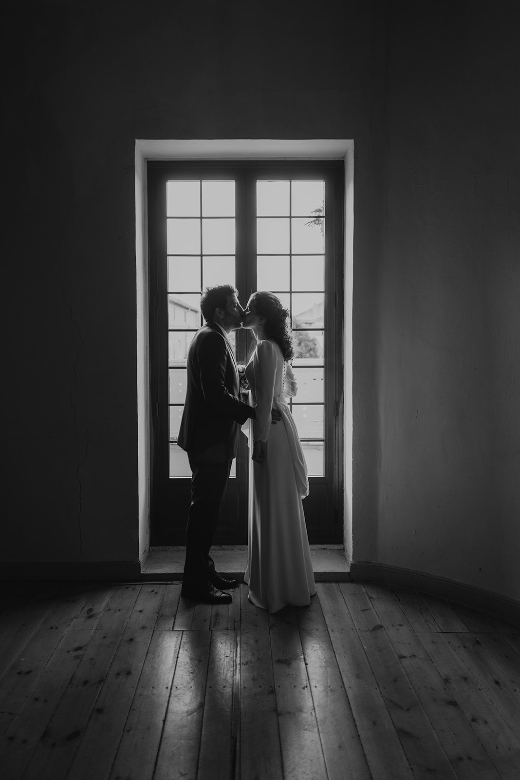 Pareja besándose de pie frente a una ventana grande, en un ambiente de luz tenue.