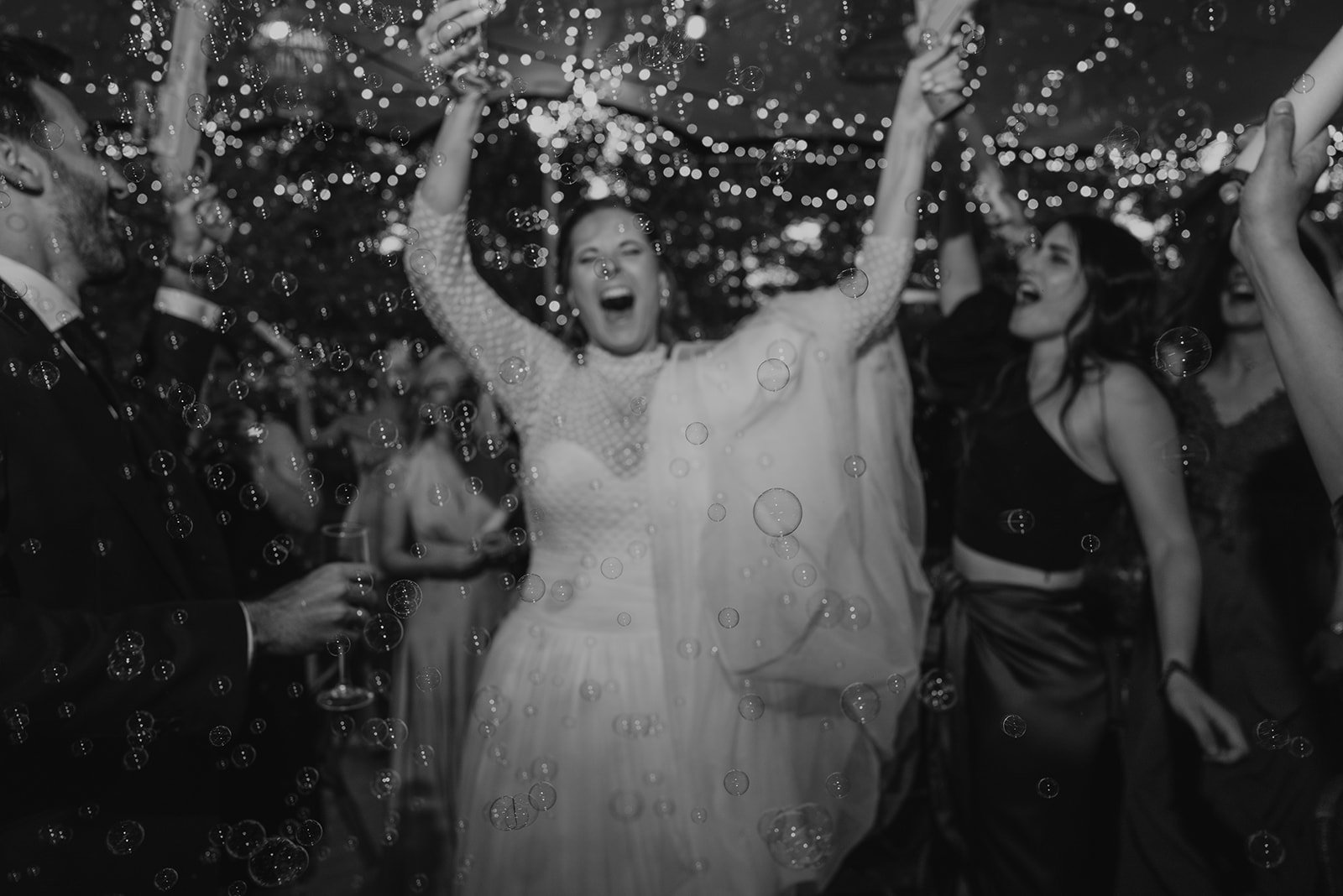 Personas celebrando en una fiesta, rodeadas de burbujas y luces brillantes, levanten copas de champán en el aire. Ambiente de celebración y alegría.