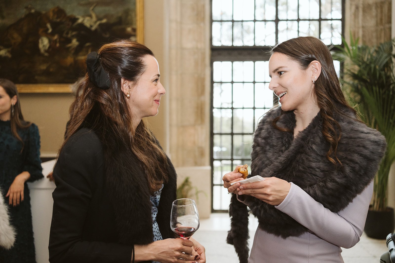 Dos mujeres conversando en un evento social, una sostiene una copa de vino tinto y la otra un aperitivo, ambas vestidas elegantemente con abrigos de piel.