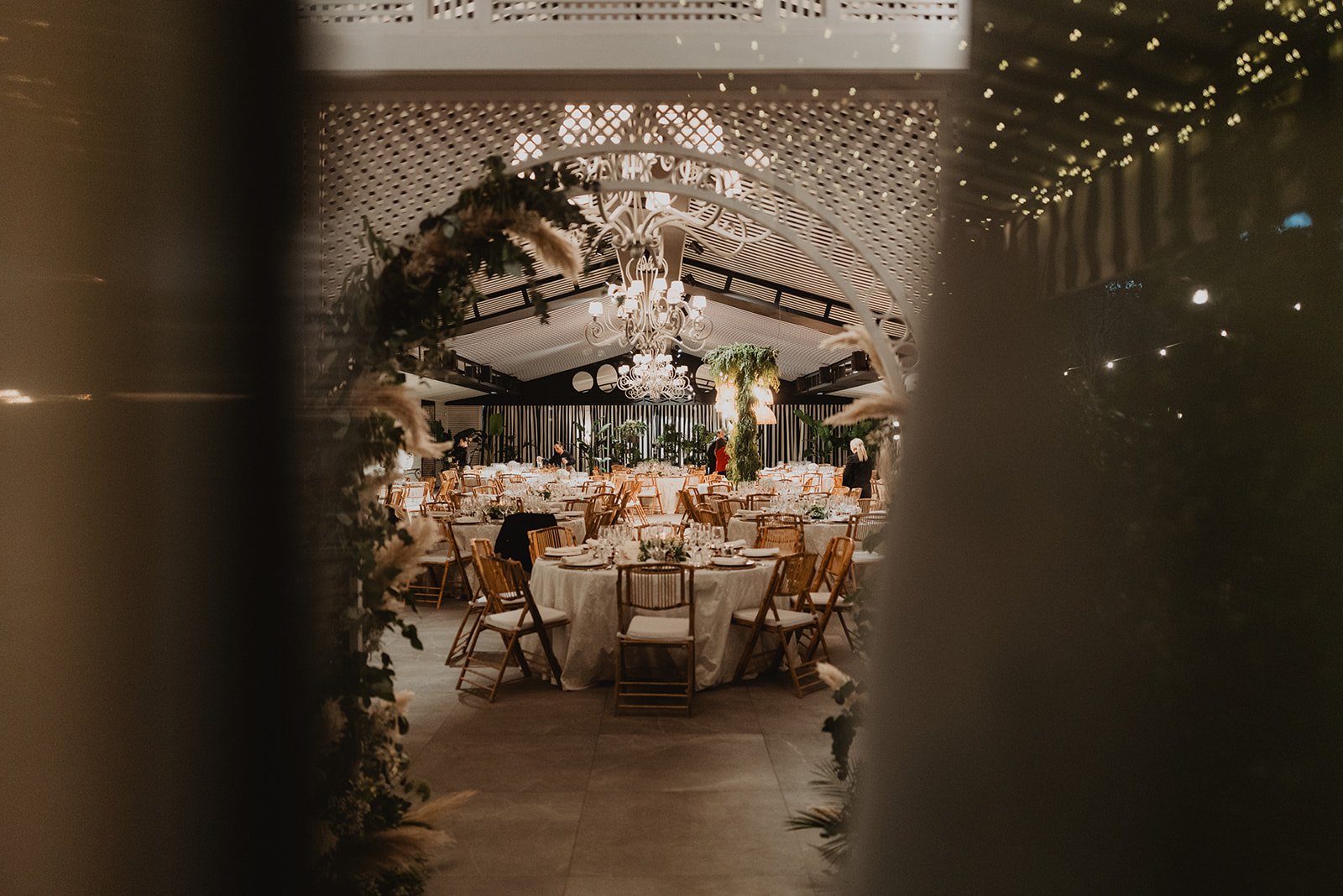 Salón de eventos decorado para una boda, con mesas redondas, sillas de madera, mantel blanco y candelabros en el techo.