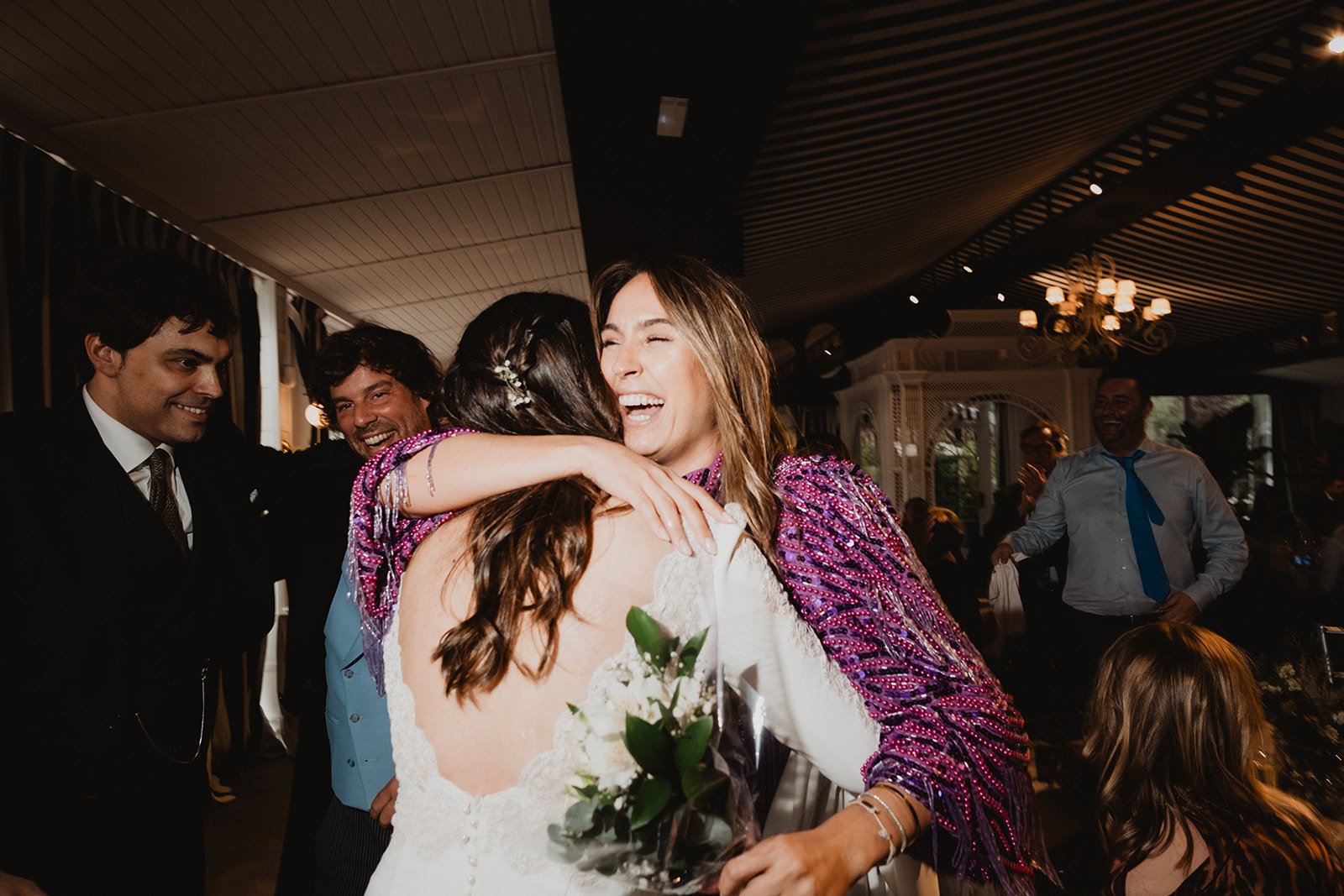Grupo de personas celebrando en una fiesta, abrazando y sonriendo. Una mujer lleva un vestido de novia con ramo de flores, mientras otra mujer, con un chal brillante, la abraza.