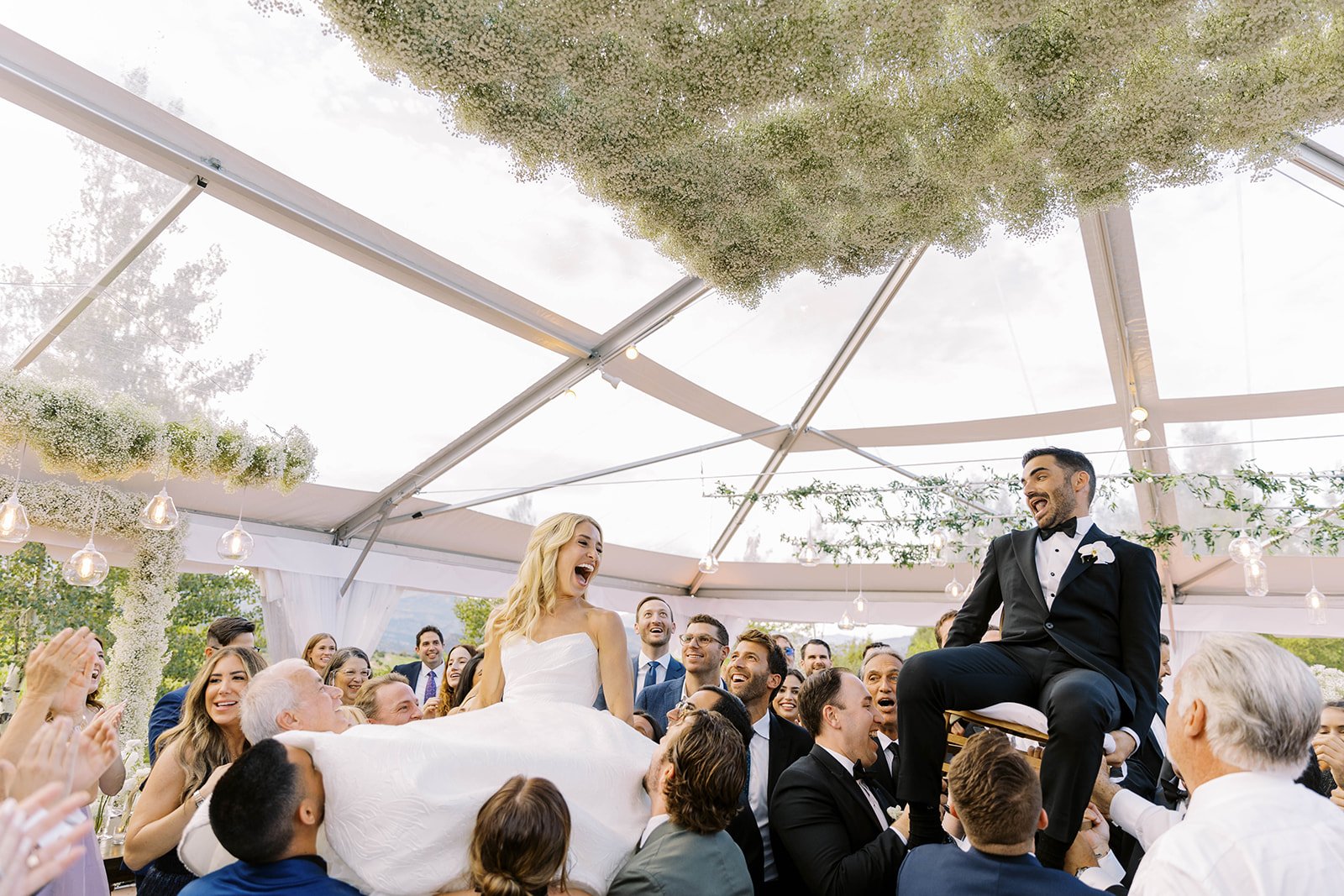 A wedding celebration with the bride and groom being lifted on chairs by guests in a decorated outdoor tent, surrounded by smiling guests.