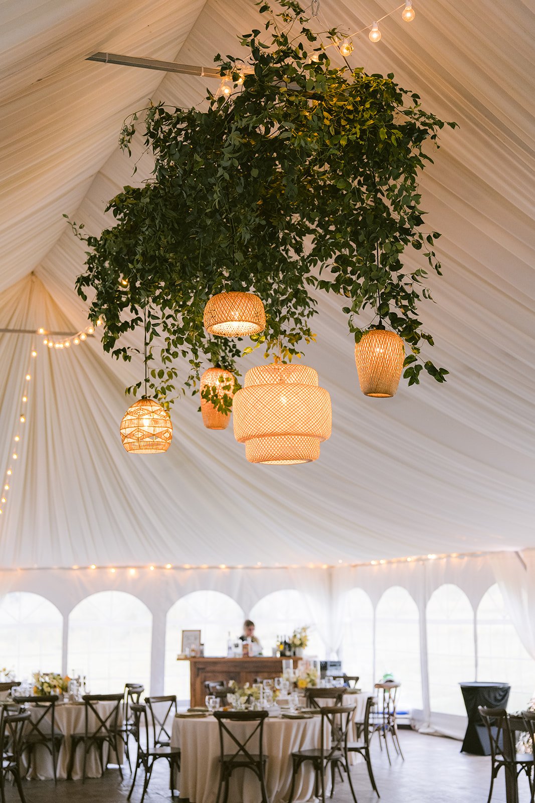 Interior of an event tent with draped ceiling, hanging greenery, wicker pendant lights, and tables set for dining. A bartender is visible at a counter in the background. Maui wedding planner.