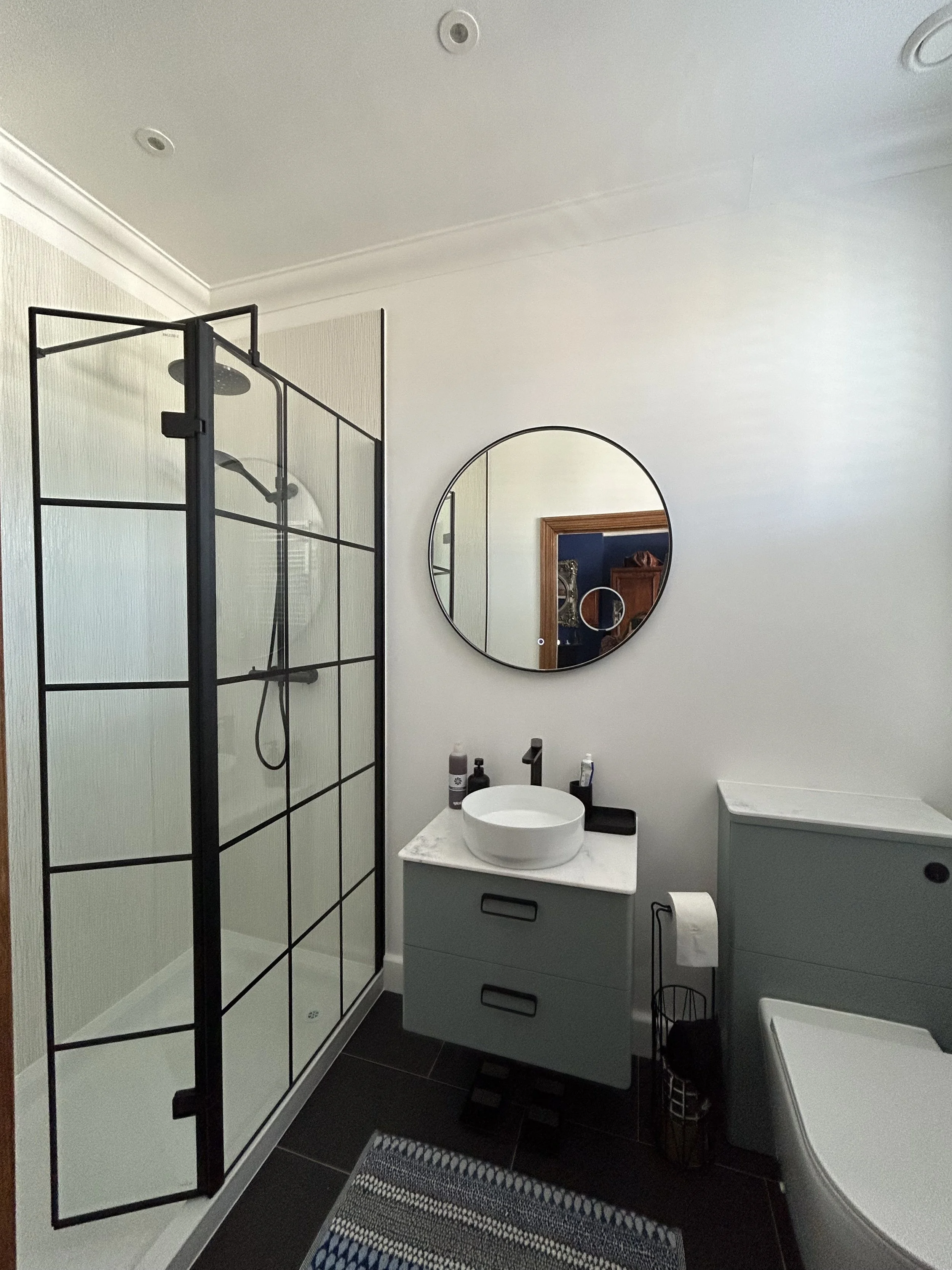 Modern bathroom with a walk-in shower with black-framed glass doors, a round mirror above a gray vanity with a vessel sink, a gray cabinet, and a toilet. There's a striped rug on the dark tile floor and personal care items on the vanity.