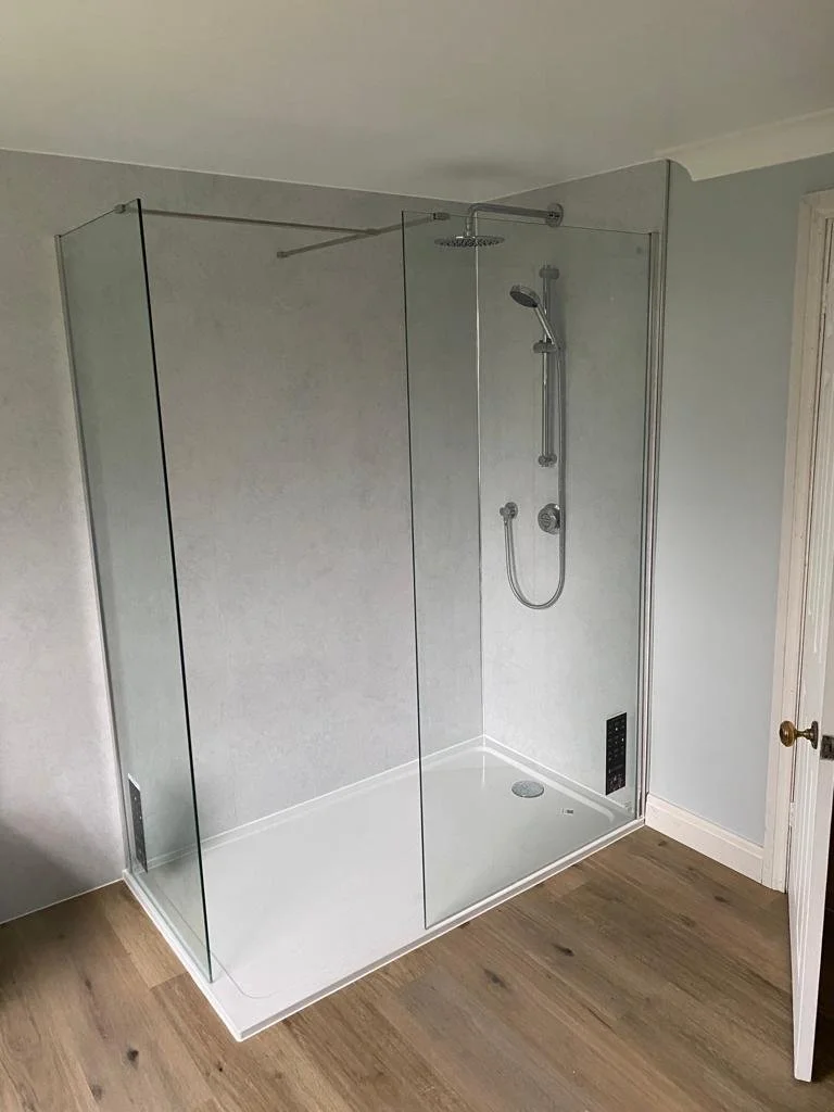 Empty walk-in shower with glass doors, wall-mounted showerhead, and handheld shower in a bathroom with wooden floor and light-colored walls.