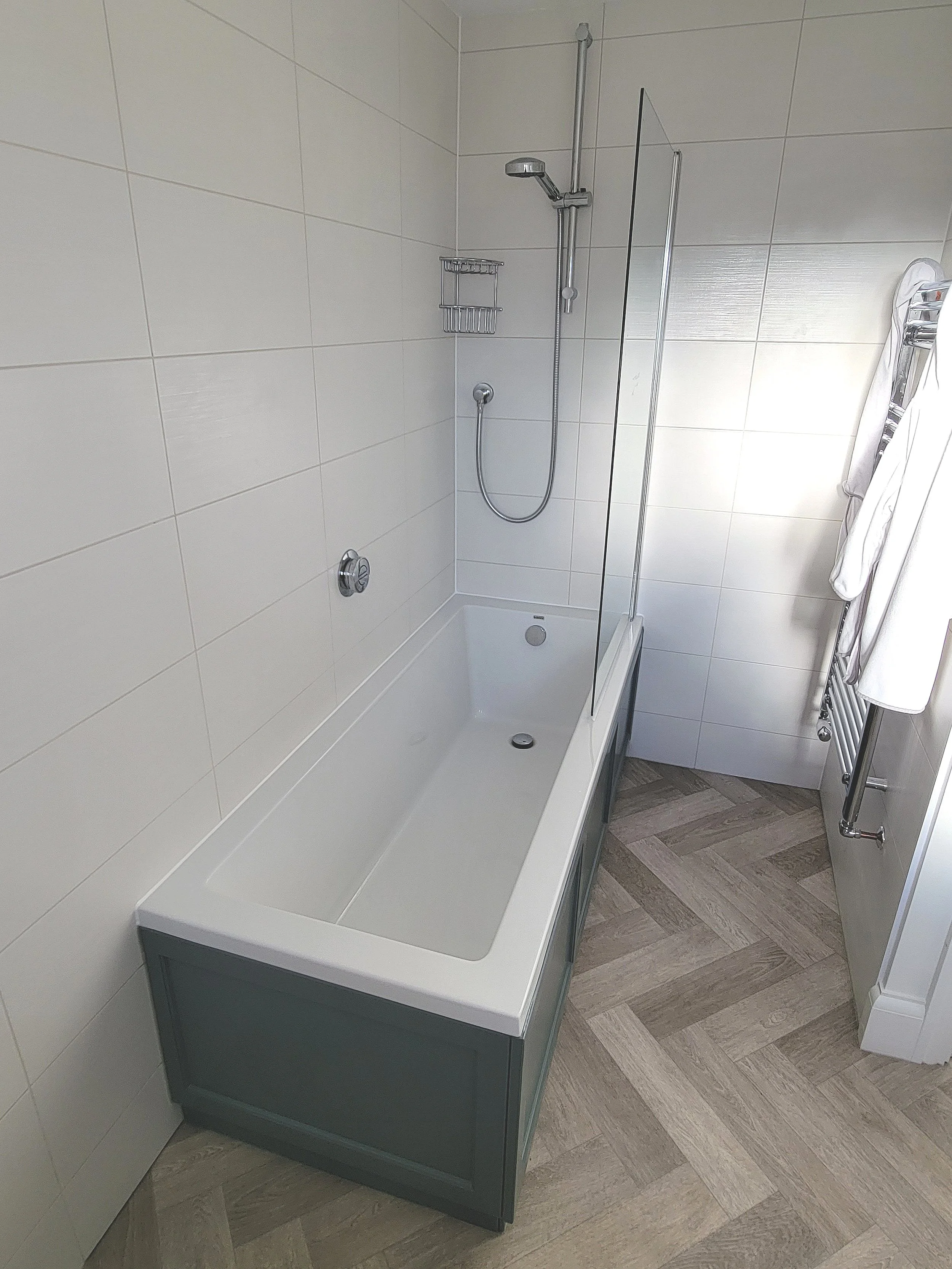 Empty bathtub in a modern bathroom with white tiled walls, a showerhead, a glass partition, and a towel rack with white towels.