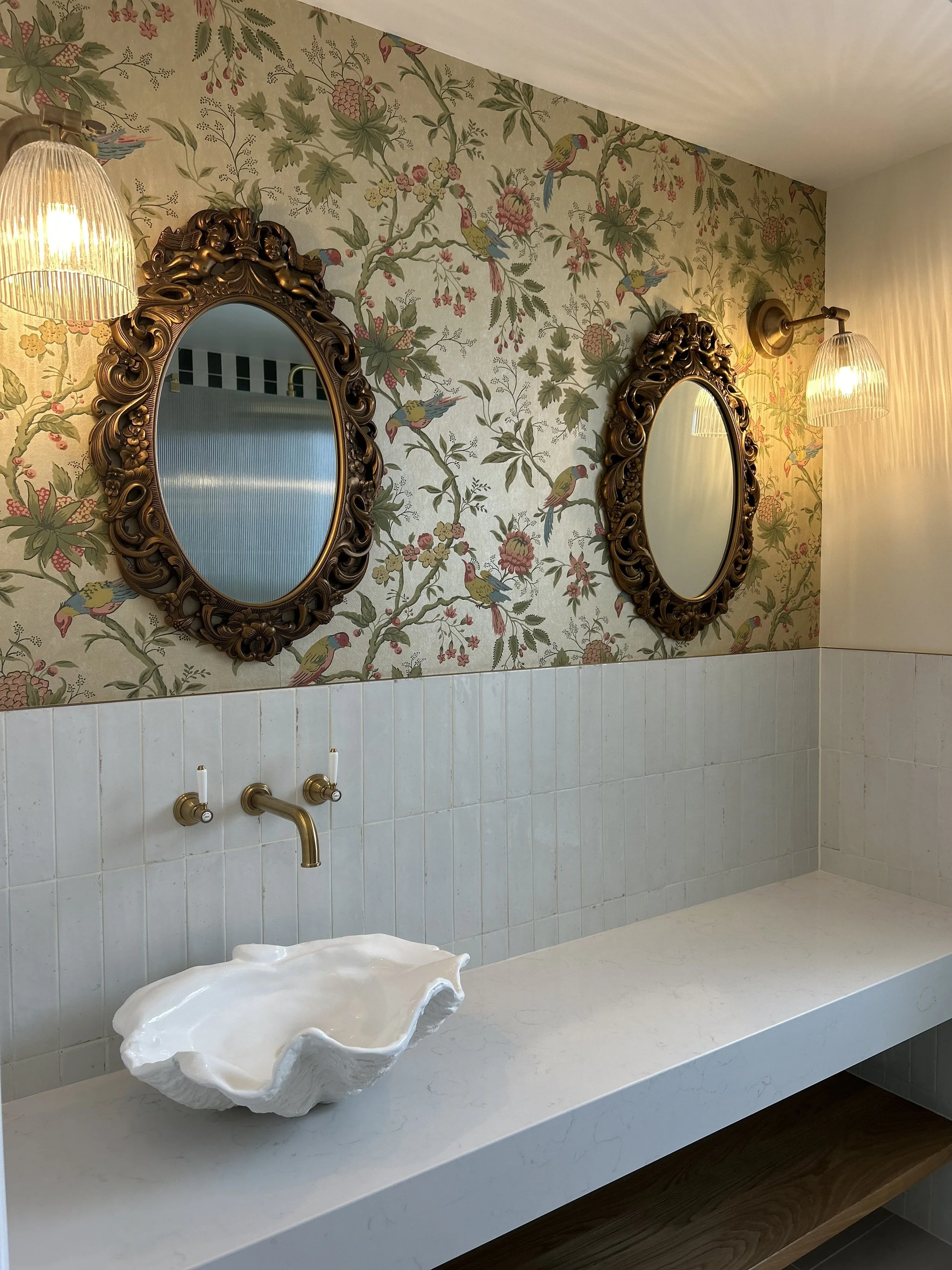 Bathroom with floral wallpaper featuring birds, two ornate oval mirrors, a shell-shaped white sink, brass faucet and lighting fixtures, and tiled lower wall with a marble countertop.