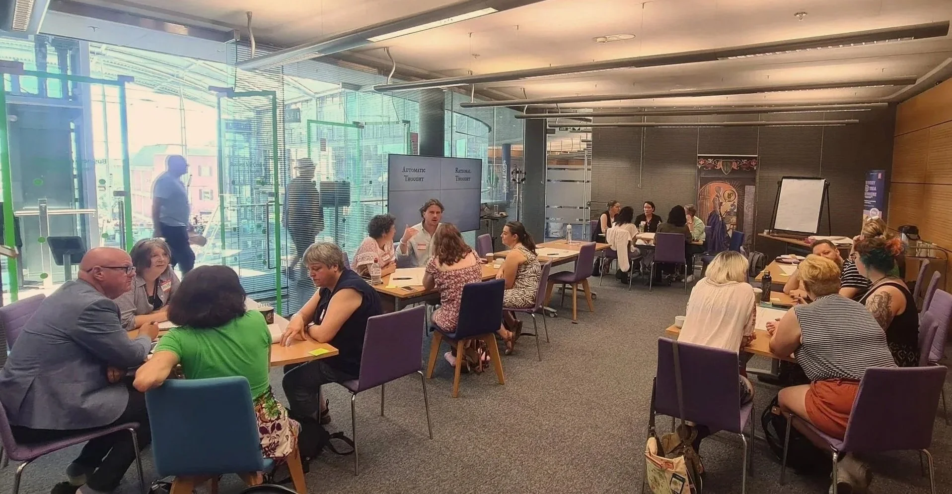 An image depicting a room of people in groups of 4 or 5 around tables. The people are discussing the topic of automatic and rational thought, using post it notes and large A0 size paper to collaboratively disucss the topic of communication.
