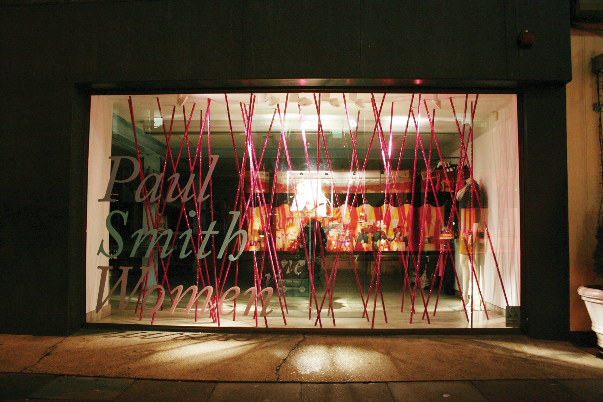 Storefront of a Paul Smith Women store with a glass window display featuring red diagonal lines and interior lights.