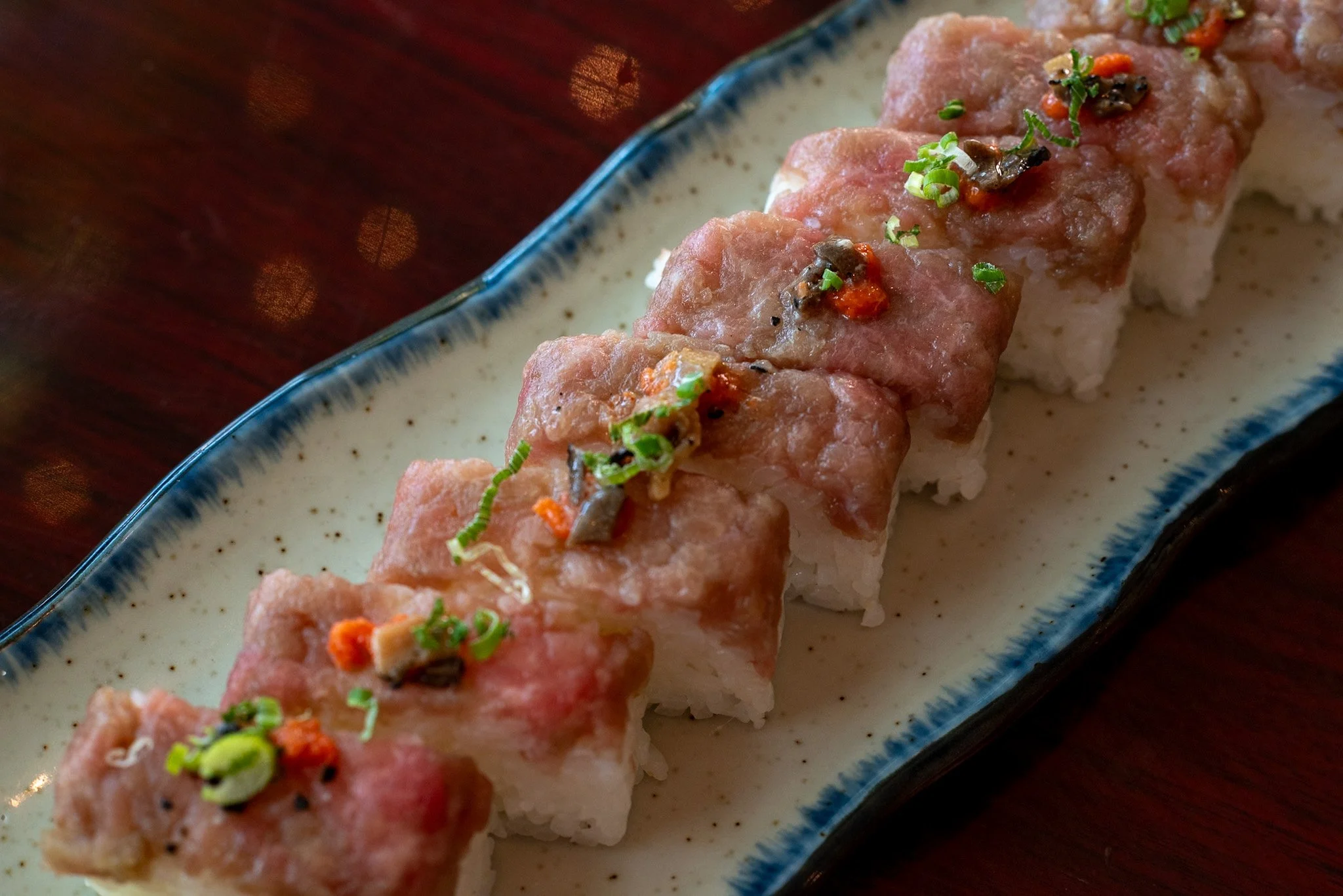 Close-up of toro box sushi roll topped with chopped green onions, and sauce, served on a white and blue ceramic plate.