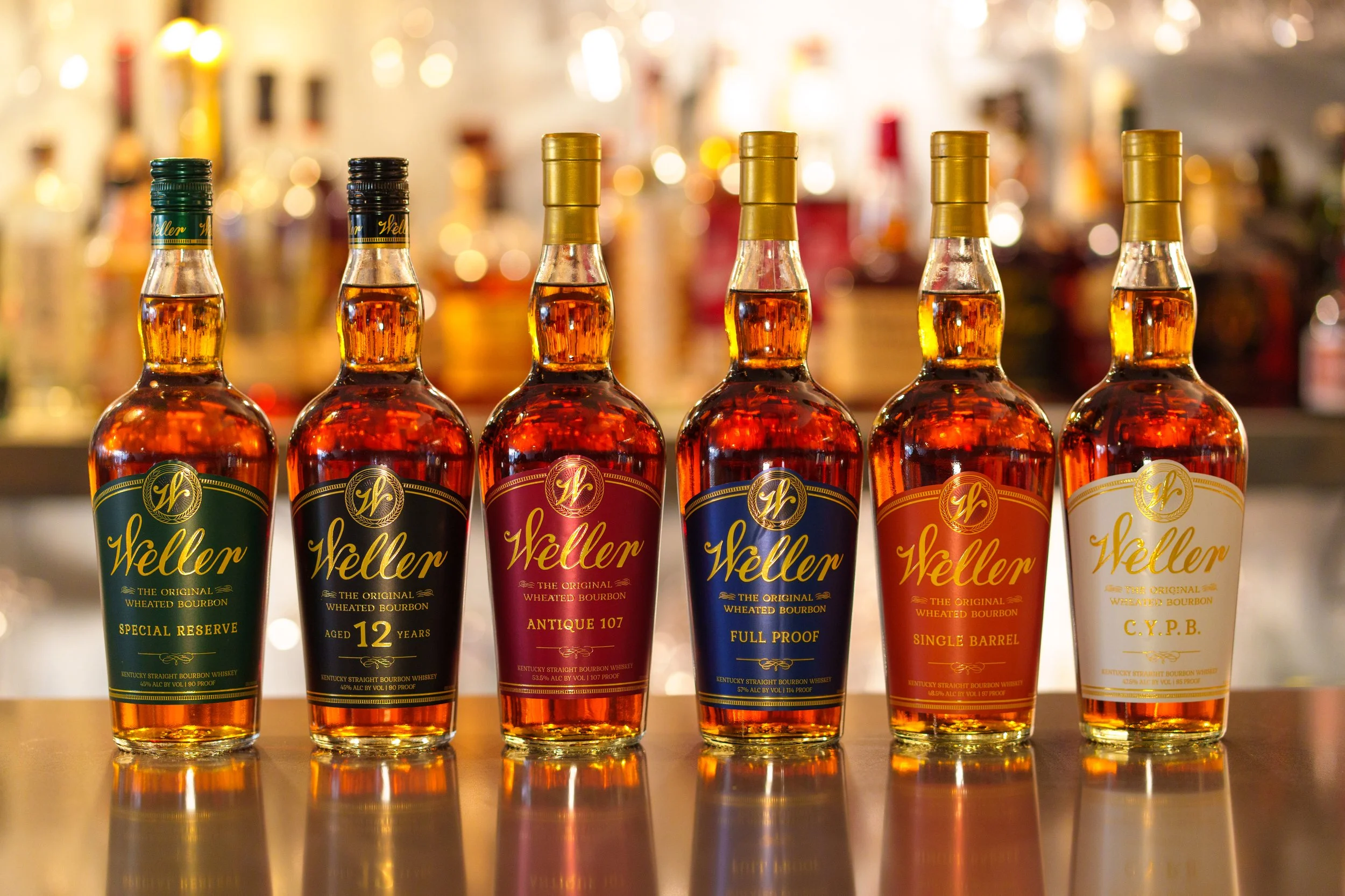 Six bottles of Weller bourbon whiskey lined up on a bar counter with a blurred background of liquor bottles and warm lighting.