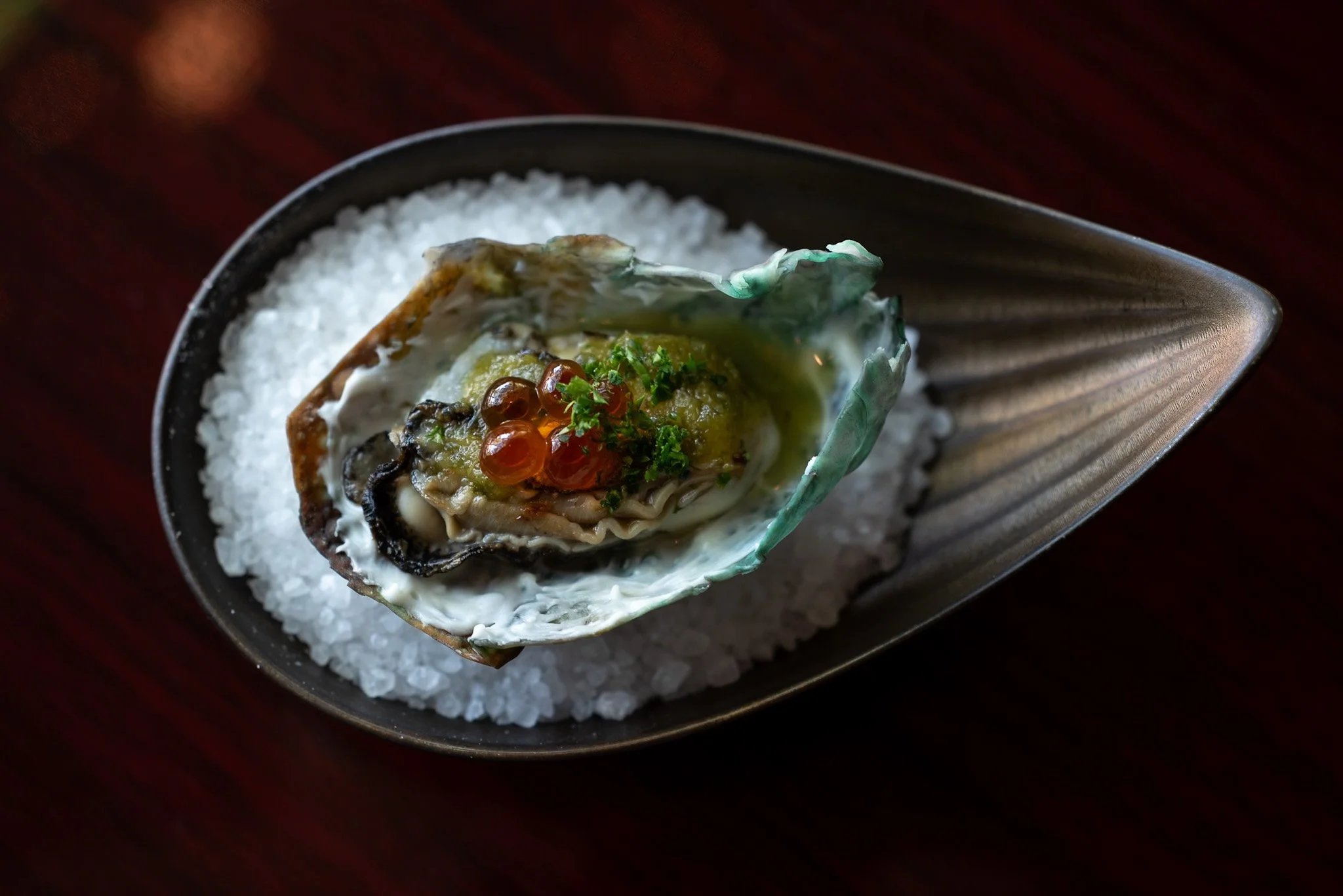 An edible oyster shell filled with oyster, topped with green herbs, red-orange roe, and white sauce, served on a bed of coarse salt in a black dish.