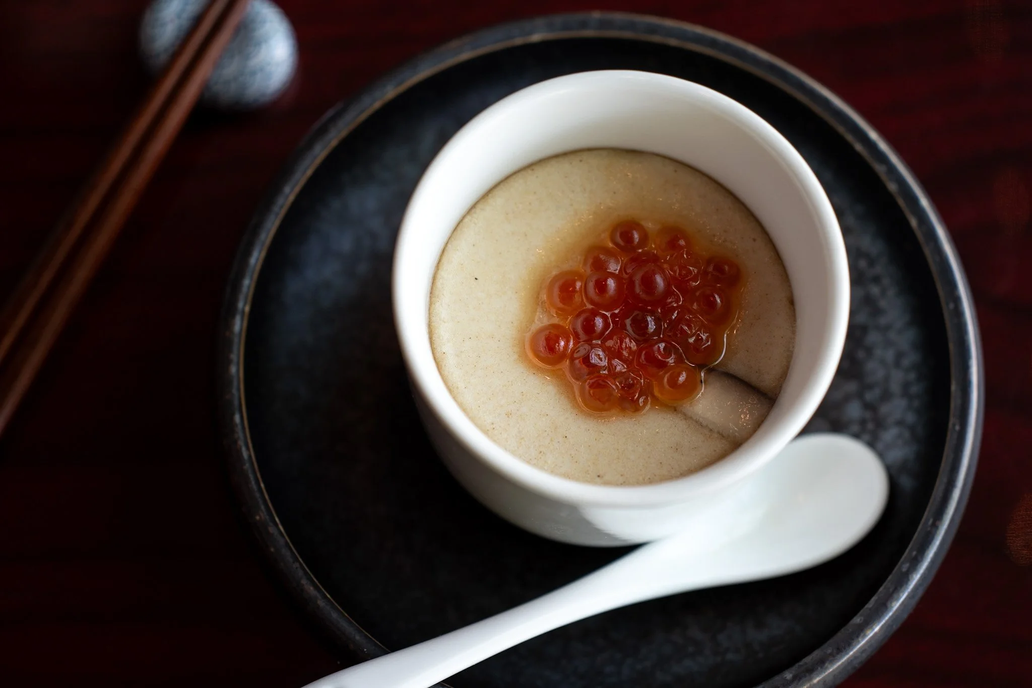 A white bowl of chawanmushi topped with red salmon roe, placed on a black plate with wooden chopsticks nearby on a dark wooden table.