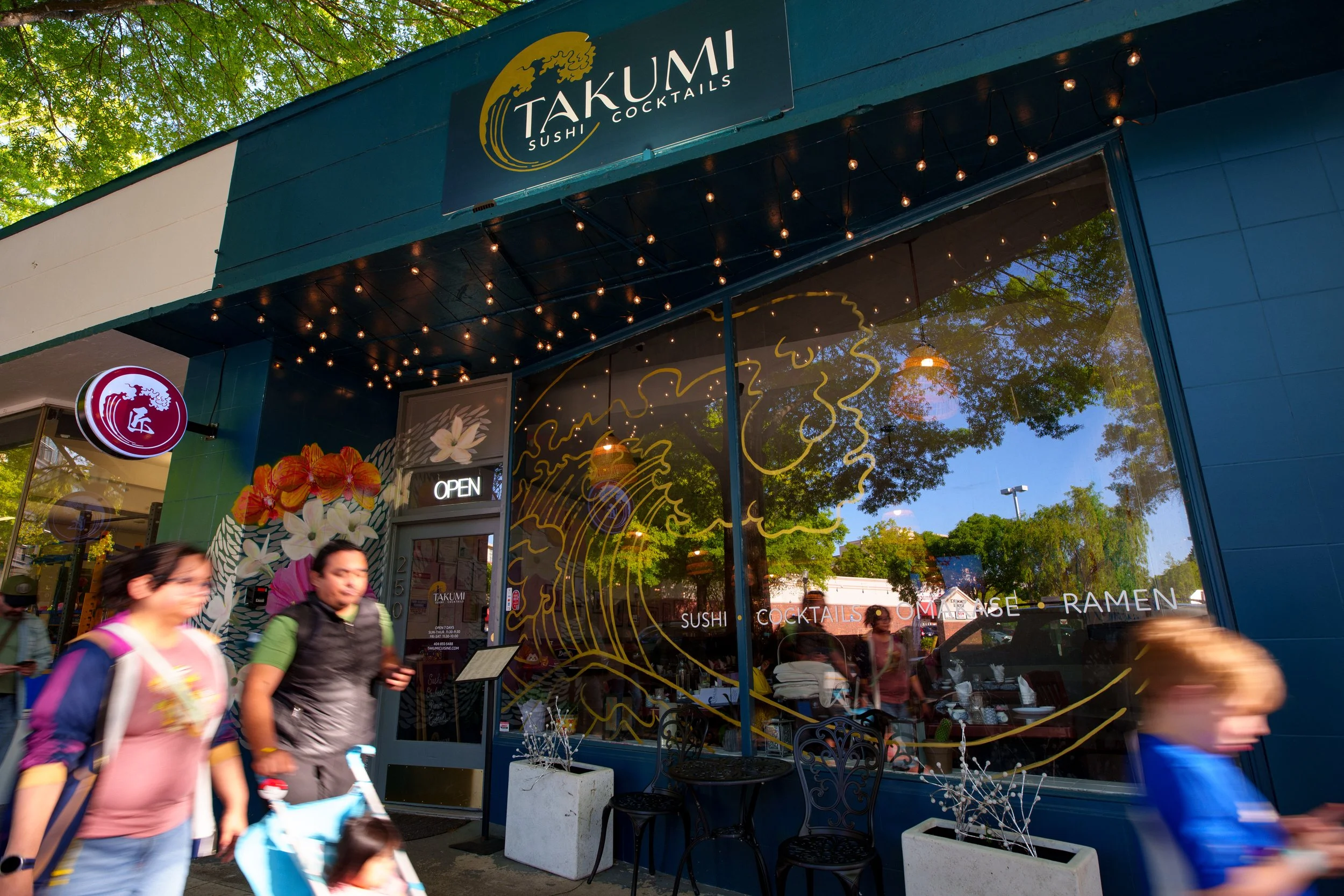 People walking past Takumi Sushi, a restaurant with a blue exterior and large window. The sign on the building reads 'Takumi Sushi Cocktails.' There are string lights hanging above the entrance and a small 'Open' sign in the window. The reflection in the window shows trees and a blue sky. The restaurant has floral decorations outside, and the scene appears to be in a busy shopping or dining area.
