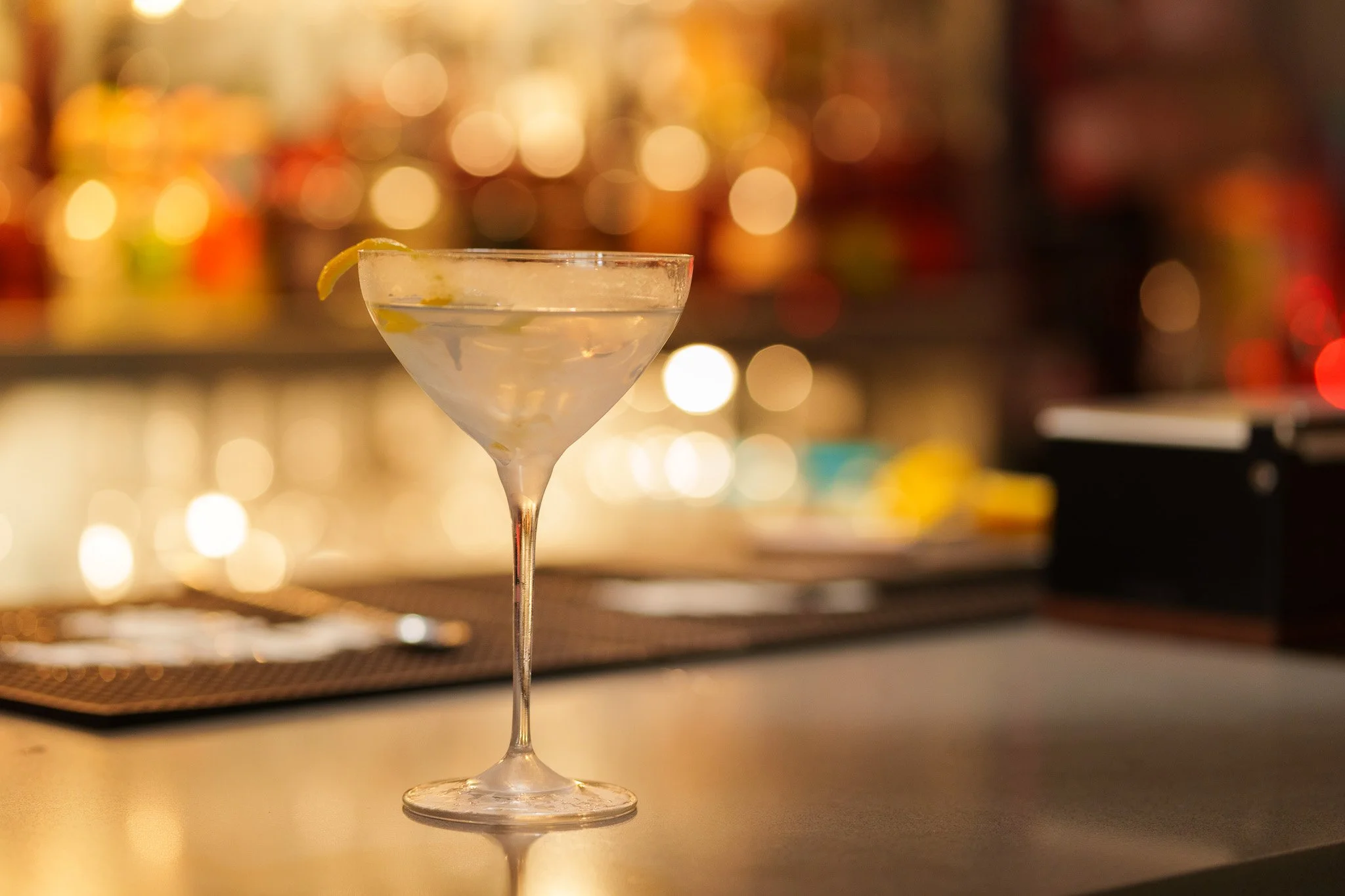 A vesper martini cocktail glass with a lemon twist garnish on a bar counter, with a blurred colorful background of bar lights.