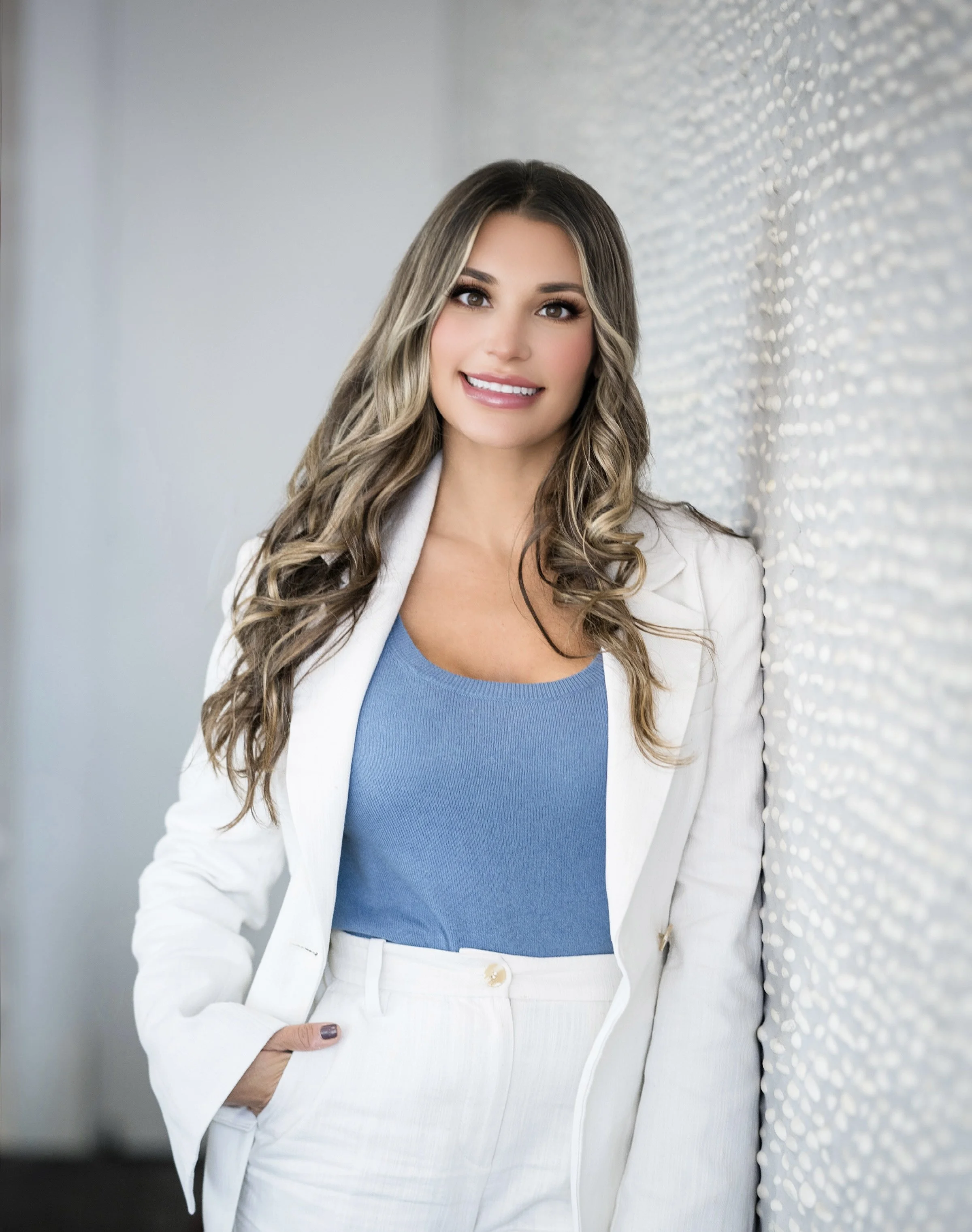 A woman with long, wavy hair wearing a white blazer and white pants, standing next to a textured wall, smiling at the camera.