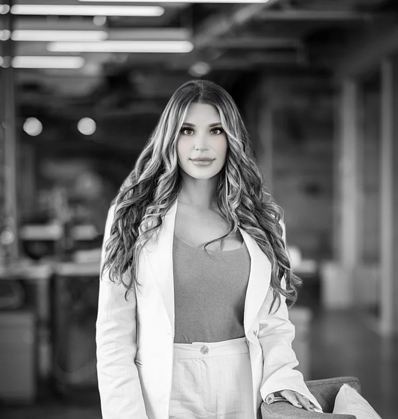 Black and white photo of a young woman with long, wavy hair, wearing a light-colored blazer over a top and high-waisted pants, standing indoors with a blurred background.