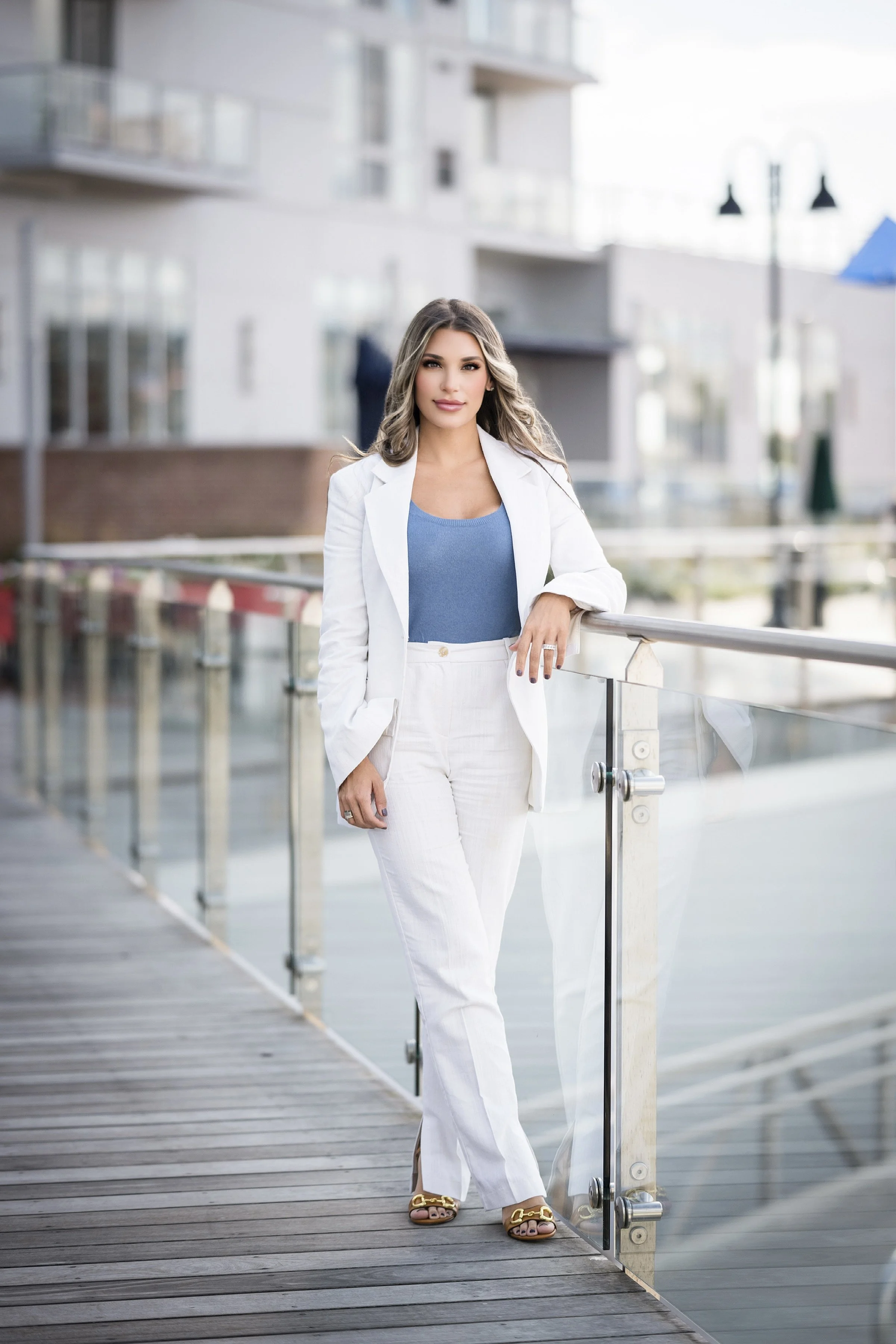 A woman with long wavy hair standing on a wooden boardwalk by water, wearing a white blazer, white pants, a blue top, and gold sandals, with buildings and street lamps in the background.